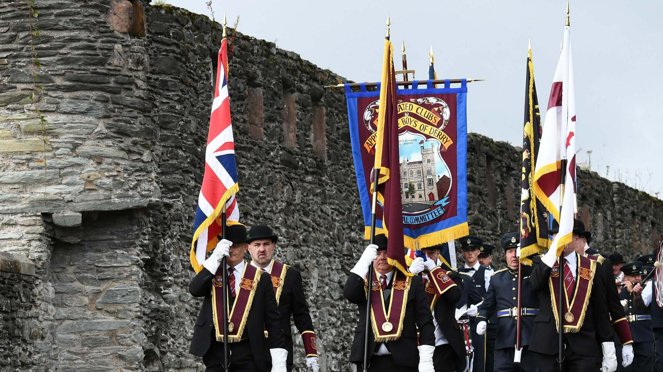 A group of men wearing black suits, black bowler hats, maroon sashes and white gloves parade next to the City of Derry's walls. There are a number of flags
