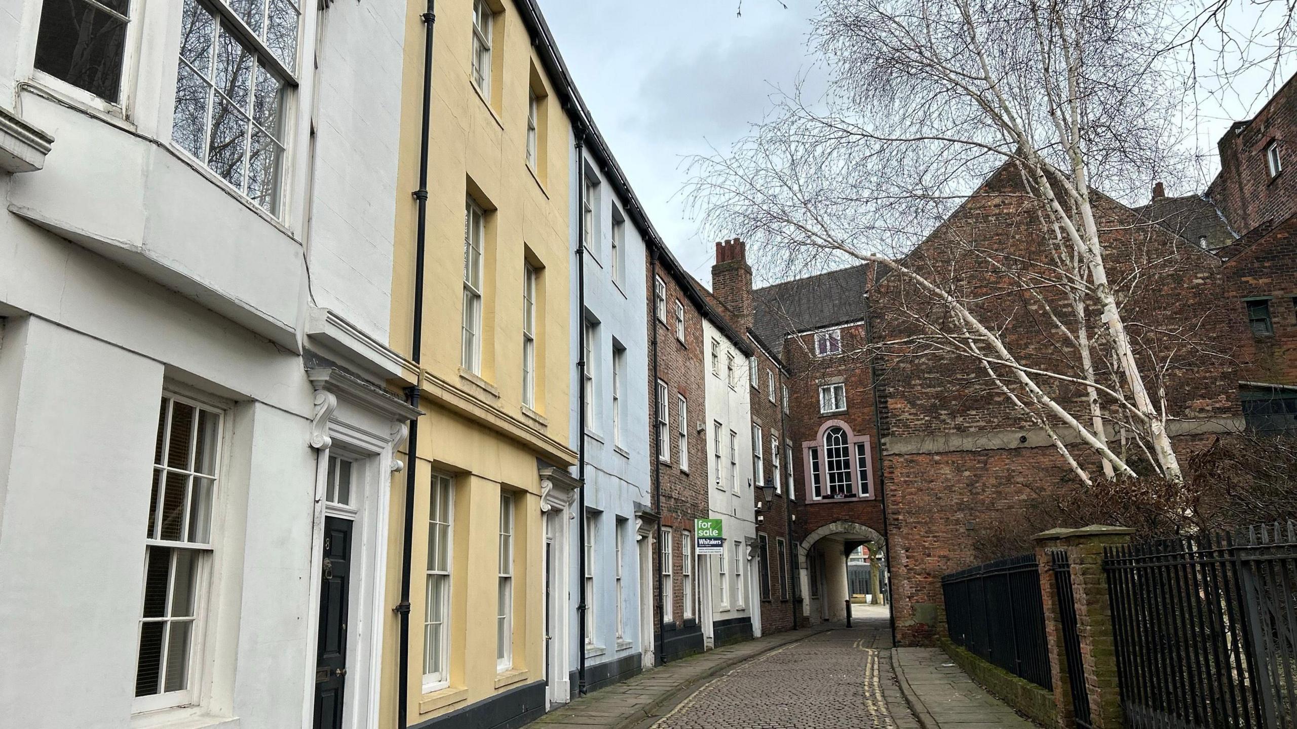 A narrow, cobbled street lined with historic townhouses painted white, yellow and pale blue. The buildings have large windows. A brick archway connects two buildings in the distance. A leafless birch tree stands behind railings on the right and the sky is blue with light clouds.
