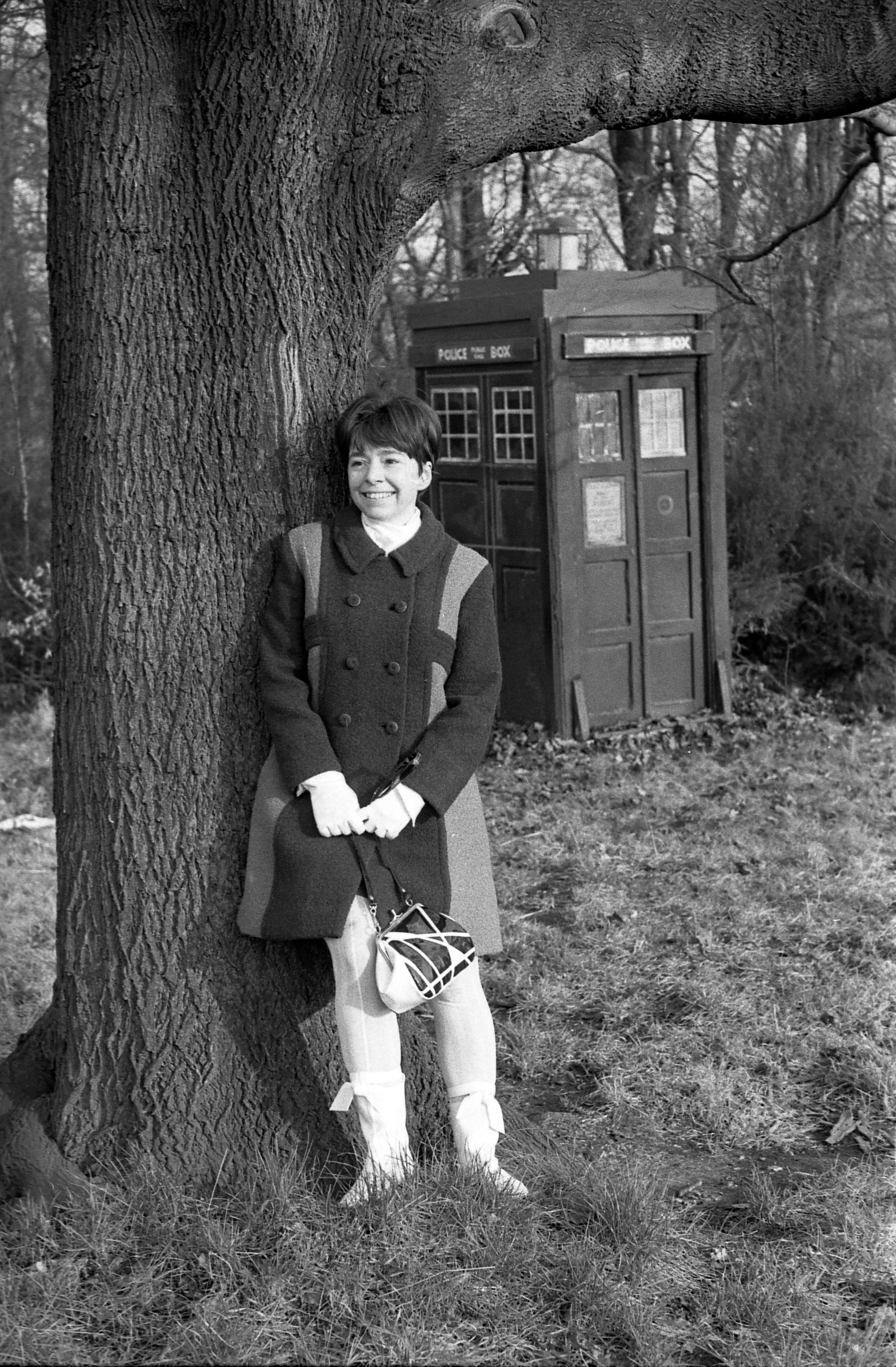 Black and white photo of Jackie Lane leaning on a large tree, with the TARDIS in the background