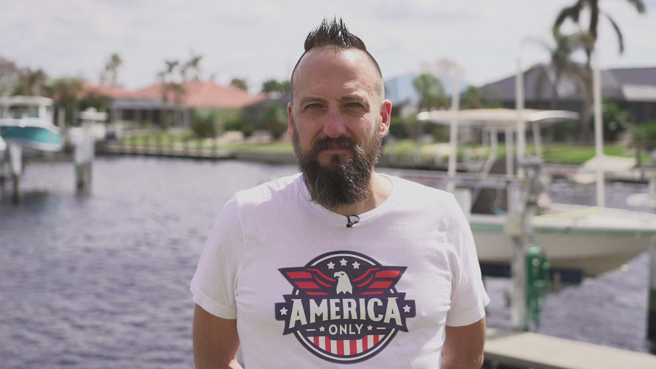 Free, a man with a full beard and closely cropped brown hair with a quiff on top, wearing a T-shirt with a stylised eagle and the slogan "America only". He is standing on a dock with boats behind him on the water