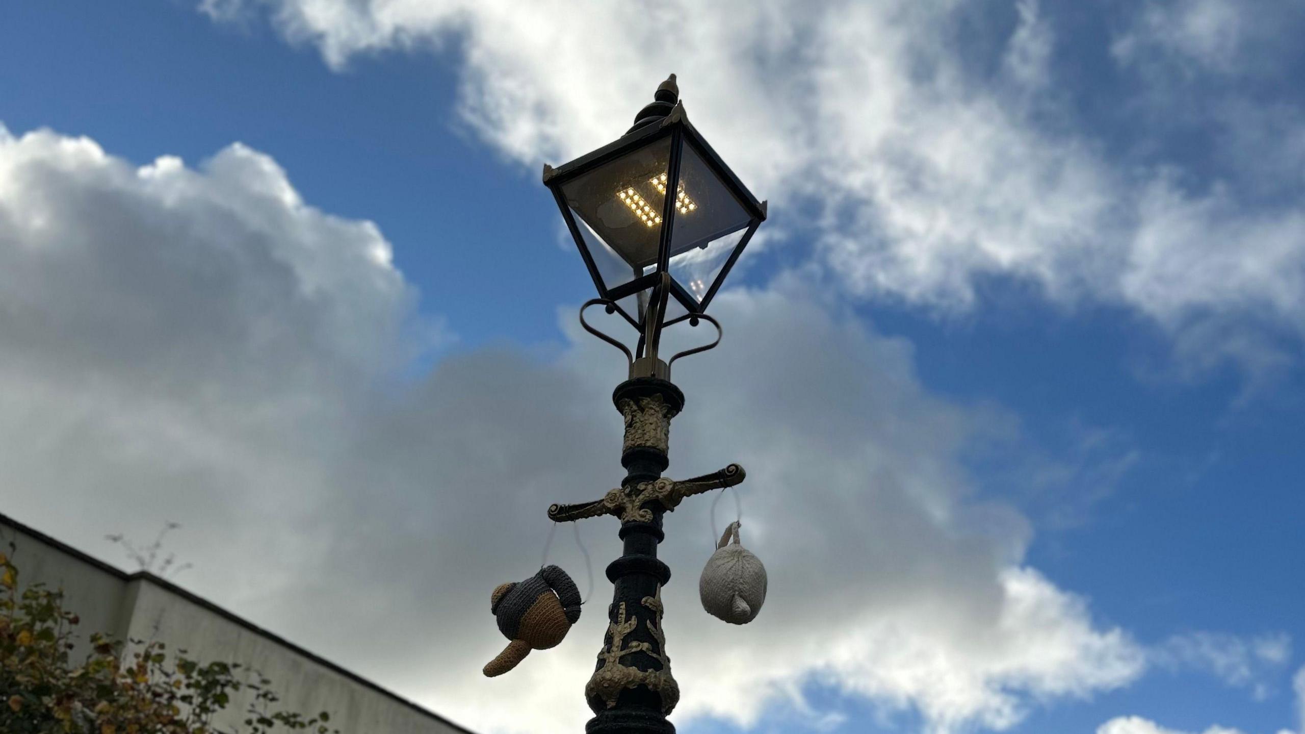 A lamppost with two knitted teapots handing from it. The sky is blue with white and grey clouds. 