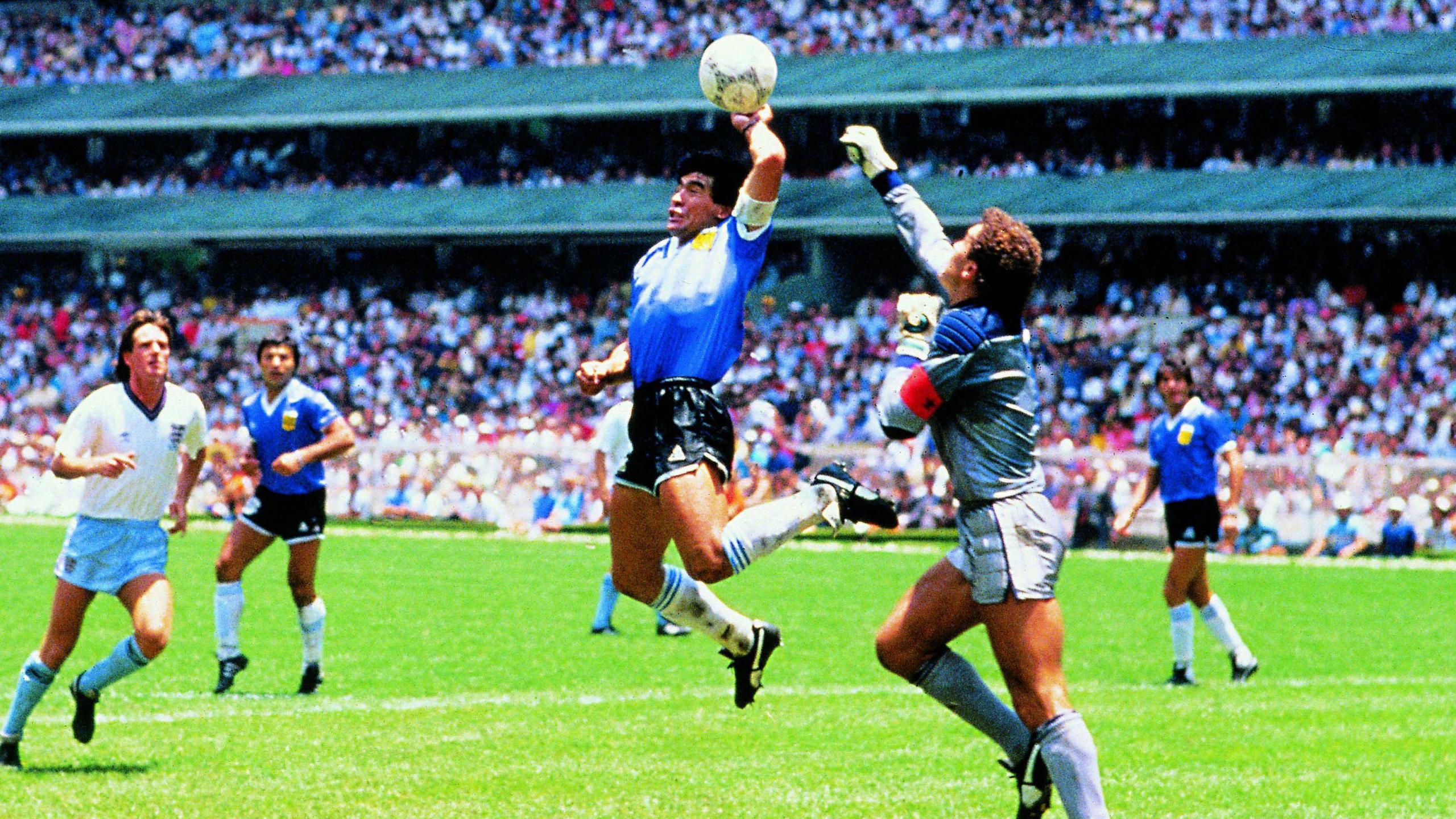 A leaping Maradona has his left hand underneath the ball as Peter Shilton attempts to punch it away. Several other players are looking on with thousands of fans in the stands behind them.