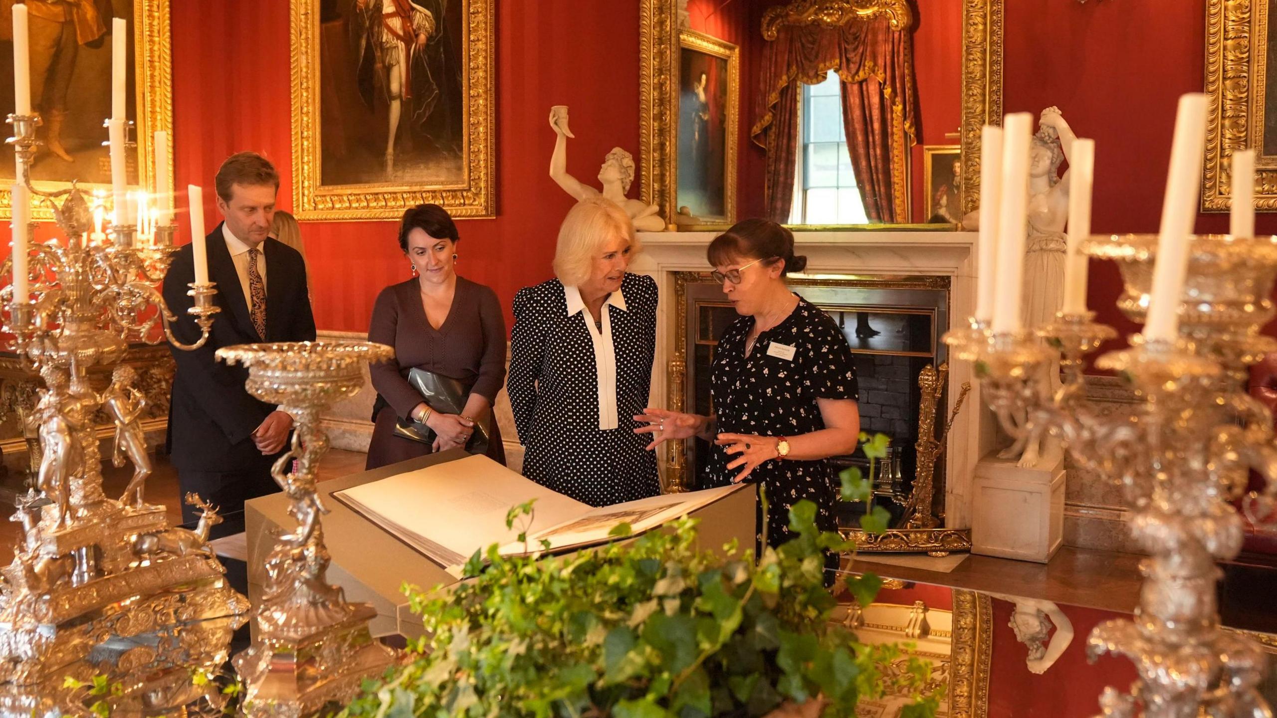 Lord Burlington (left), and Queen's Reading Room CEO Vicki Perrin (second left), watch as Queen Camilla views a selection of artefacts from the Jane Austen House museum, marking the 250th anniversary the author's birth, during The Queen's Reading Room Festival at Chatsworth House. They are in a stately room draped in red with candles on the table and old portraits on the wall.