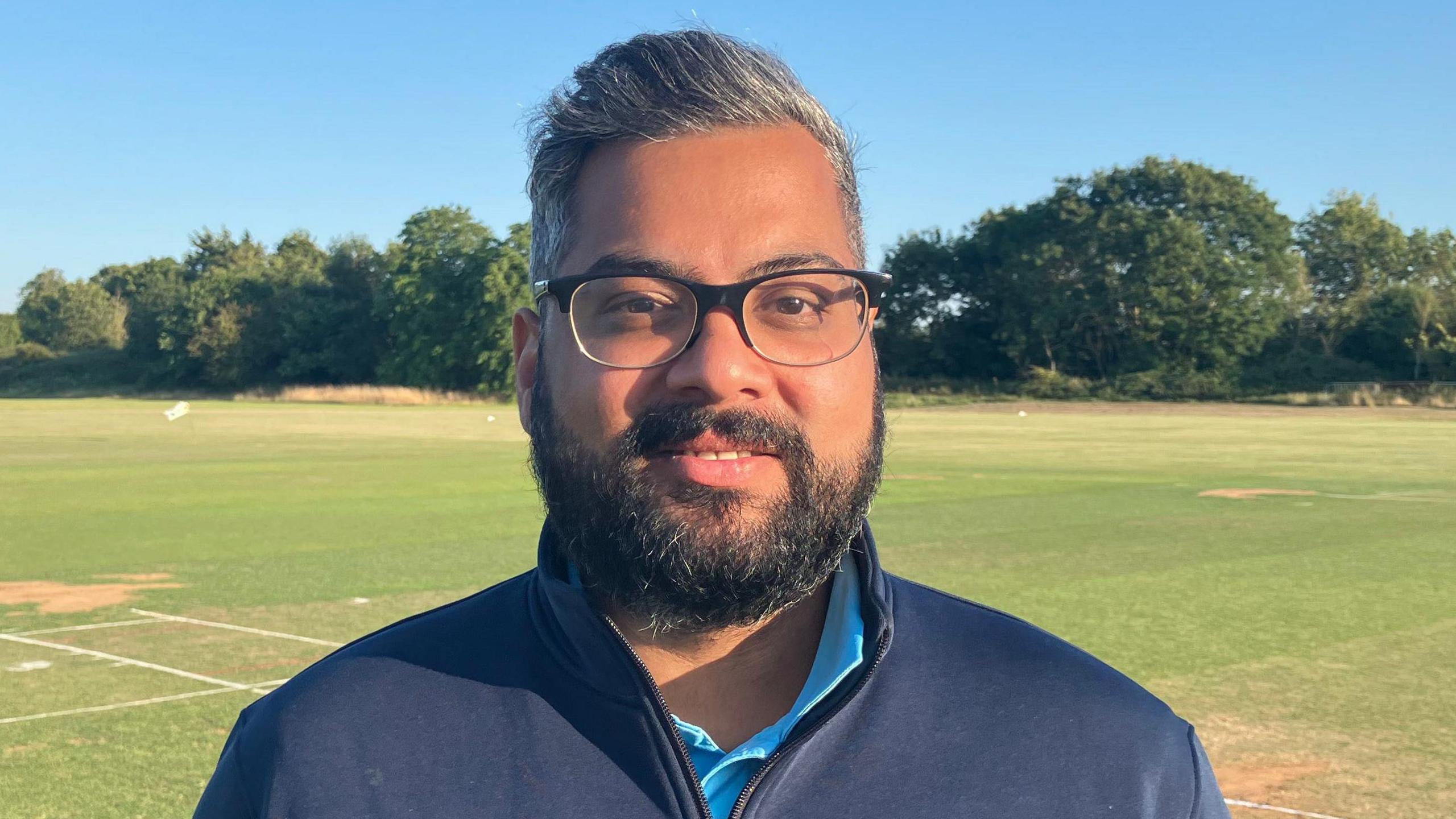 Devesh Patel is standing on the green grounds on a sunny day with blue skies. He is looking directly at the camera and smiling. He is wearing black glasses and a 3/4 zip navy jumper. He has a dark beard and short grey hair.