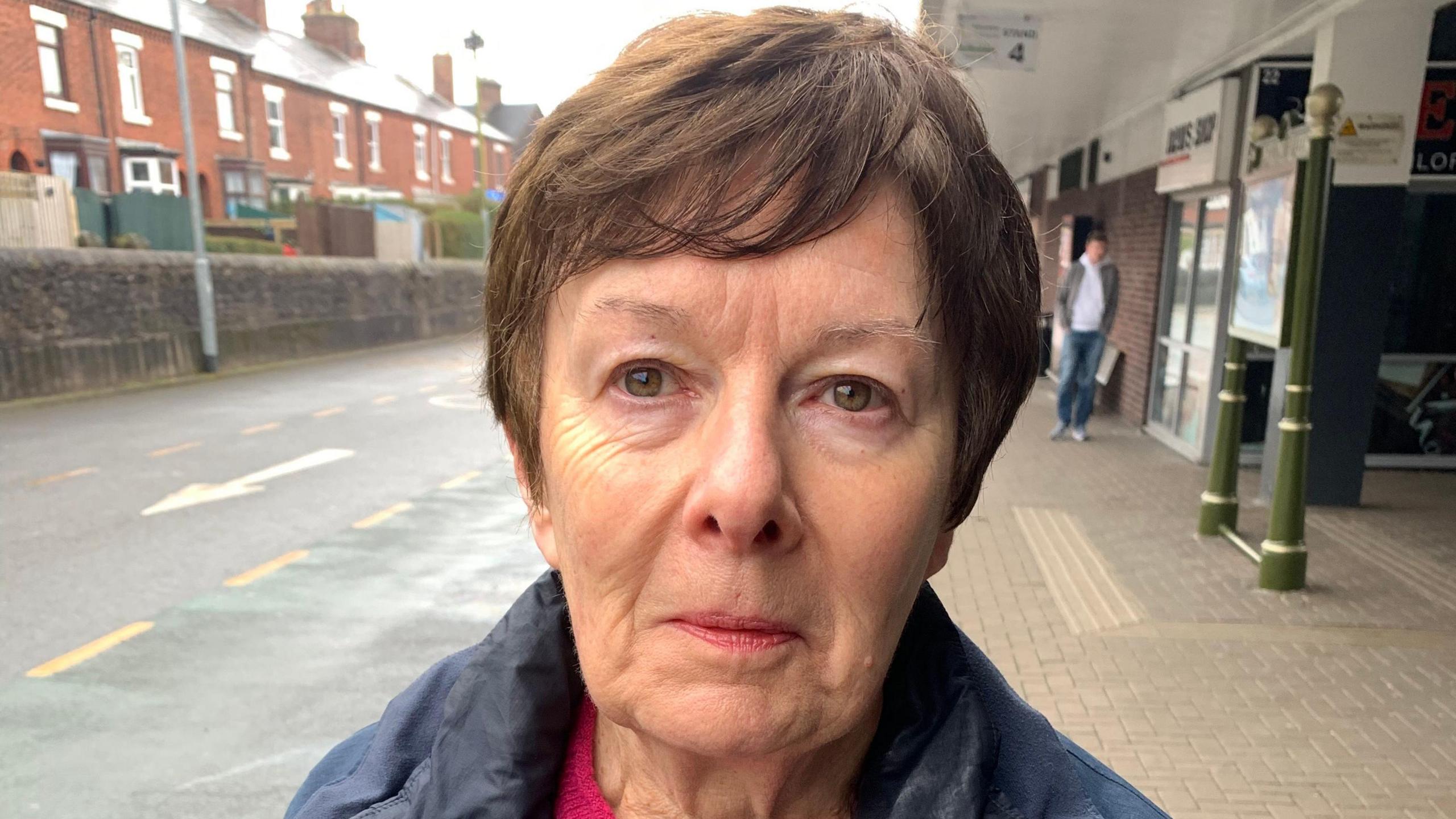 Councillor Charlotte Atkins pictured with short brown hair with a neutral expression on her face. She is standing in the middle of a street with a man in the far distance behind her, he is wearing blue jeans, a grey hoodie and jacket.  