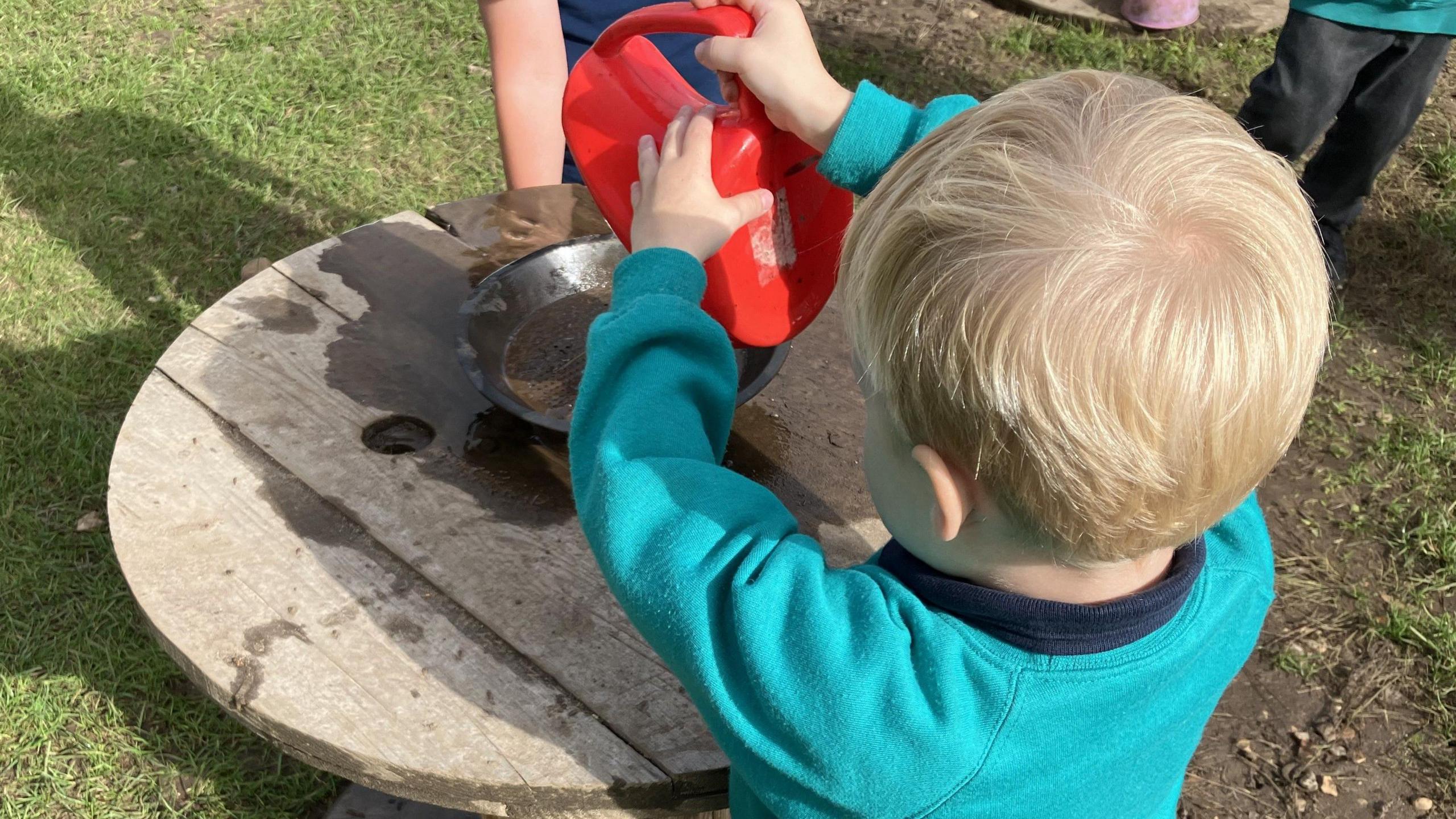 A young child is holding a red watering can and pouring water onto a silver metal dish containing dirt