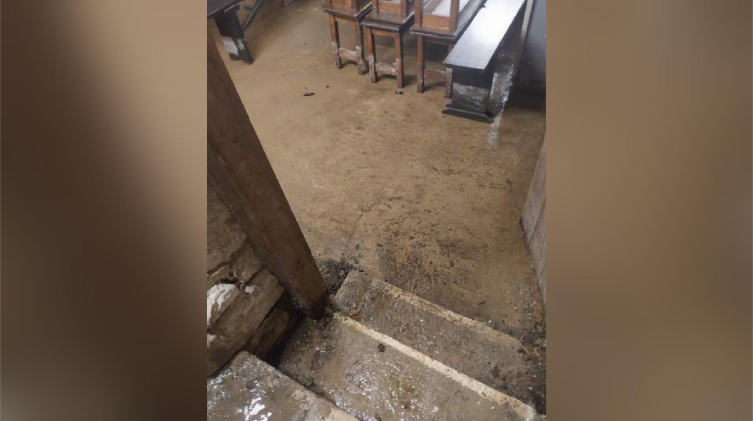 Stone steps leading down to a stone-floored room in which wooden benches are visible. There is water on the floor.