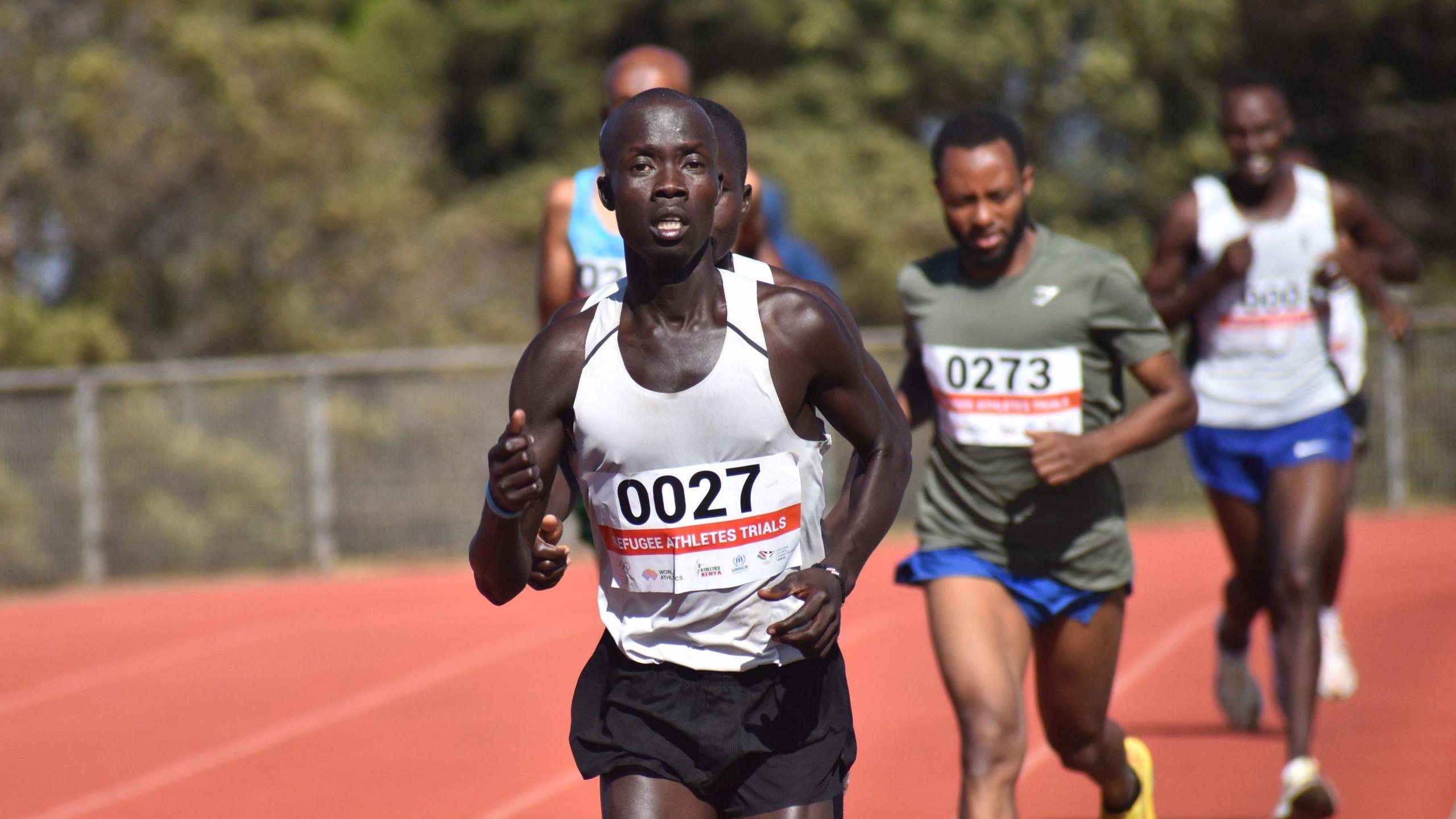 James Lokidichi, wearing a white running vest with the number 27 on it, is seen during a track race with five other men out of focus in the distance behind him