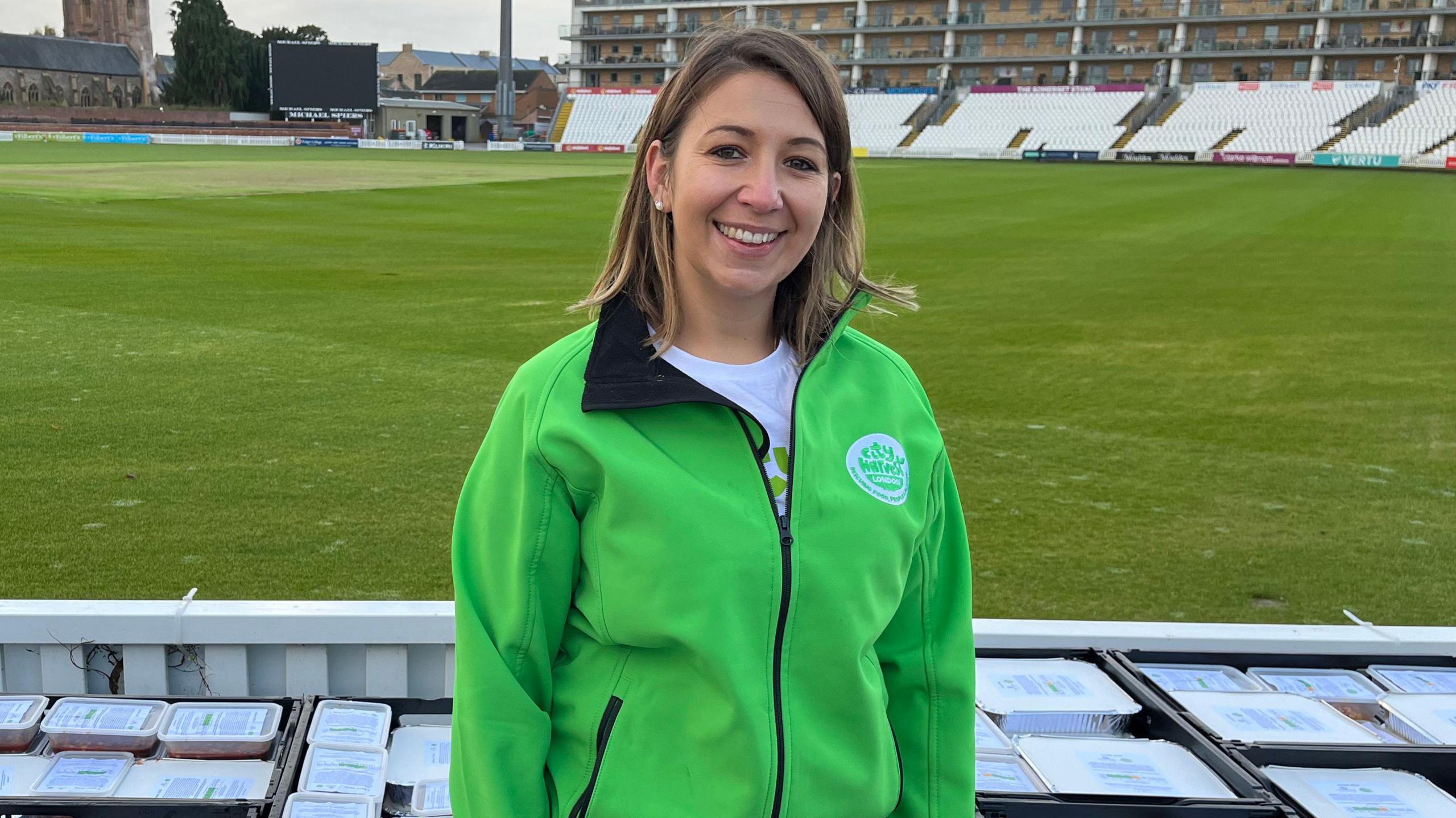 Mary Parsons is in the centre of the frame, wearing a green fleece jacket, surrounded by crates of food ready to be distributed. in the background is the cricket pitch and stands at Somerset County Cricket Club.