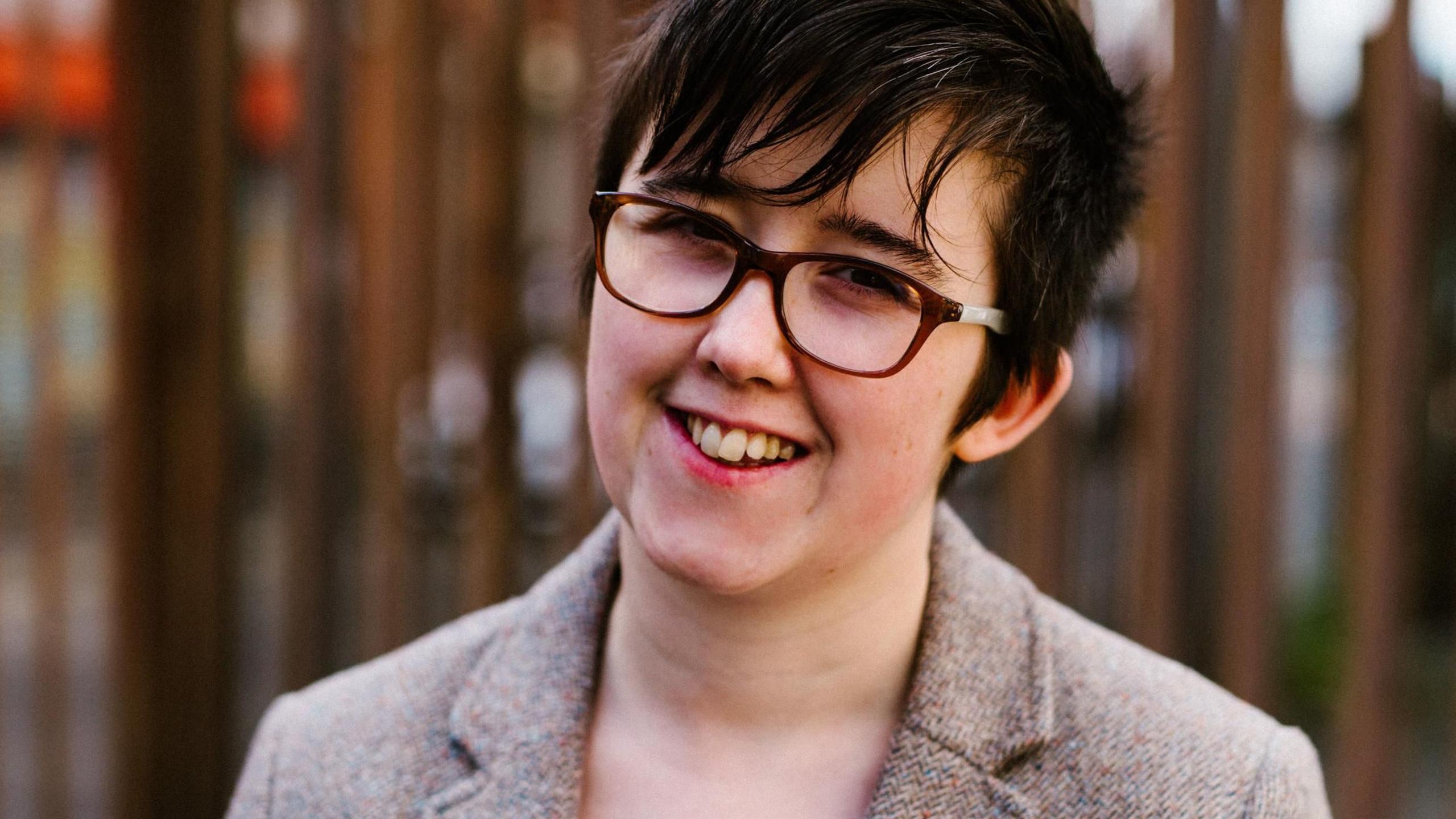 A head shot of Lyra Mckee , who was a northern ireland journalist shot dead during a riot in 2019. Wearing dark-framed glasses and a tweed suit jacket, she is smiling at the camera.