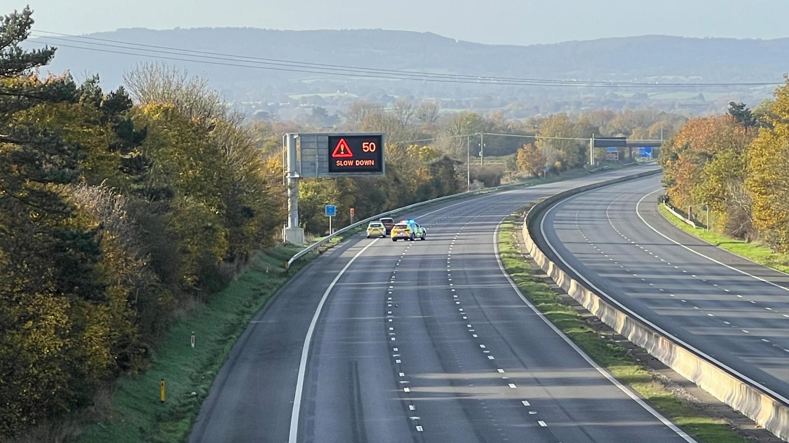 A view of an empty M5 motorway, with a "50mph slow down" matrix sign active. There are two police cars parked to one side of the road with a third dark coloured car in front of them.