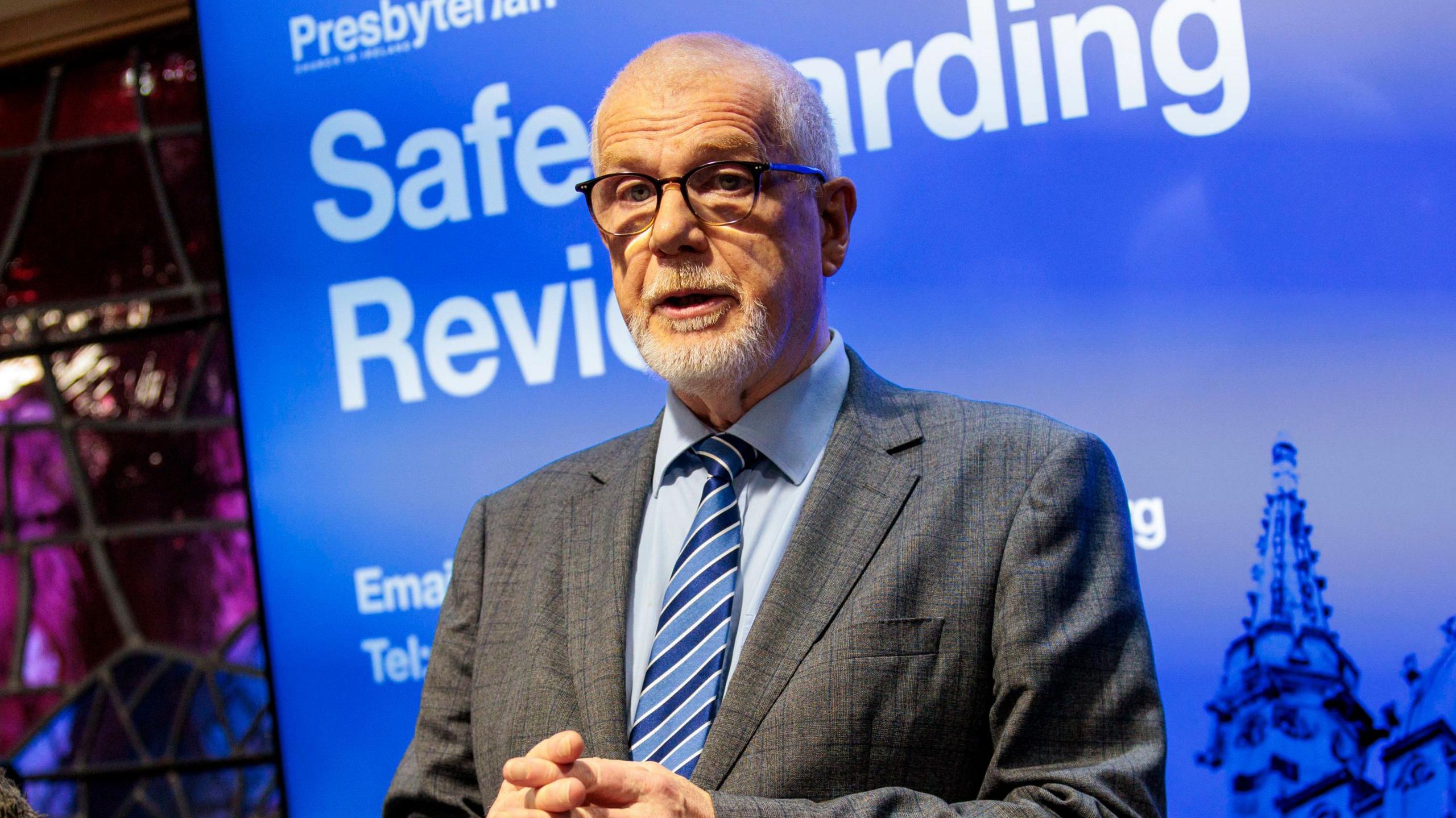 Rev David Bruce, with short grey hair and grey stubble, wearing a blue shirt, stripy blue tie and grey suit. He is standing in front of a large screen with a blue background. There is white text on the screen reading "Safeguarding Review".