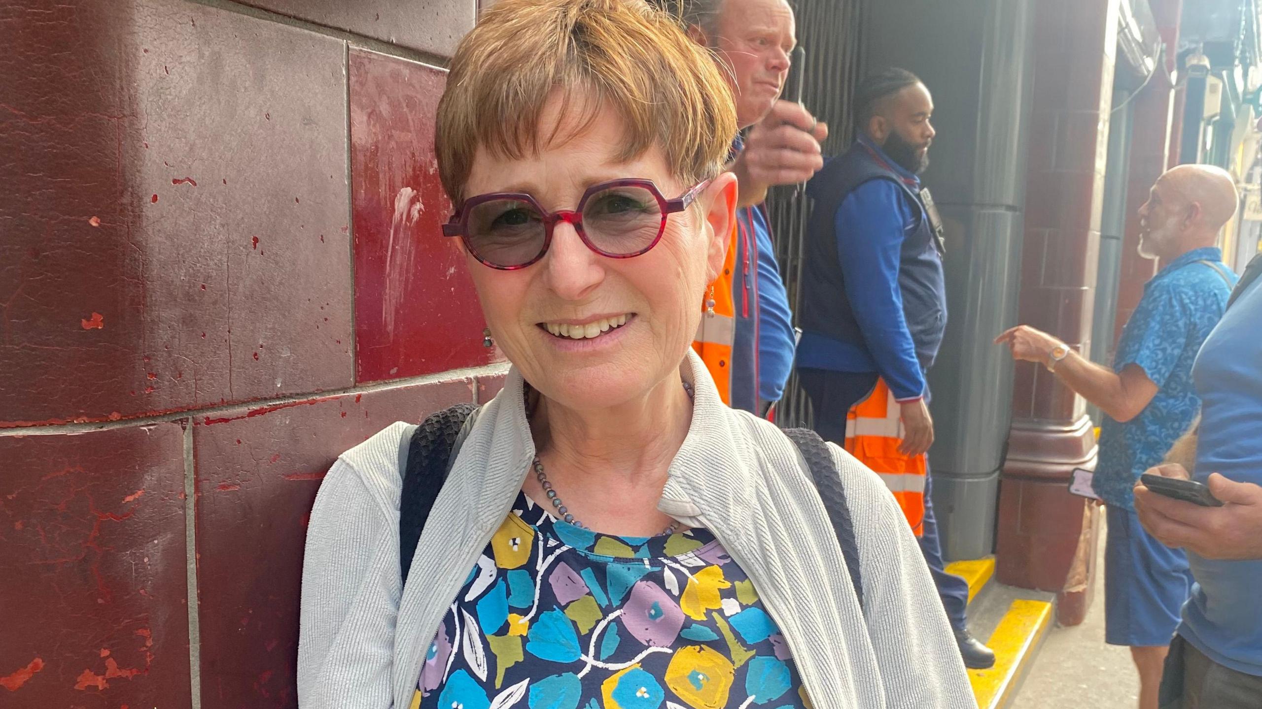 Smiling woman in colourful floral top and sunglasses stands against a red tiled wall outside a London Underground station.