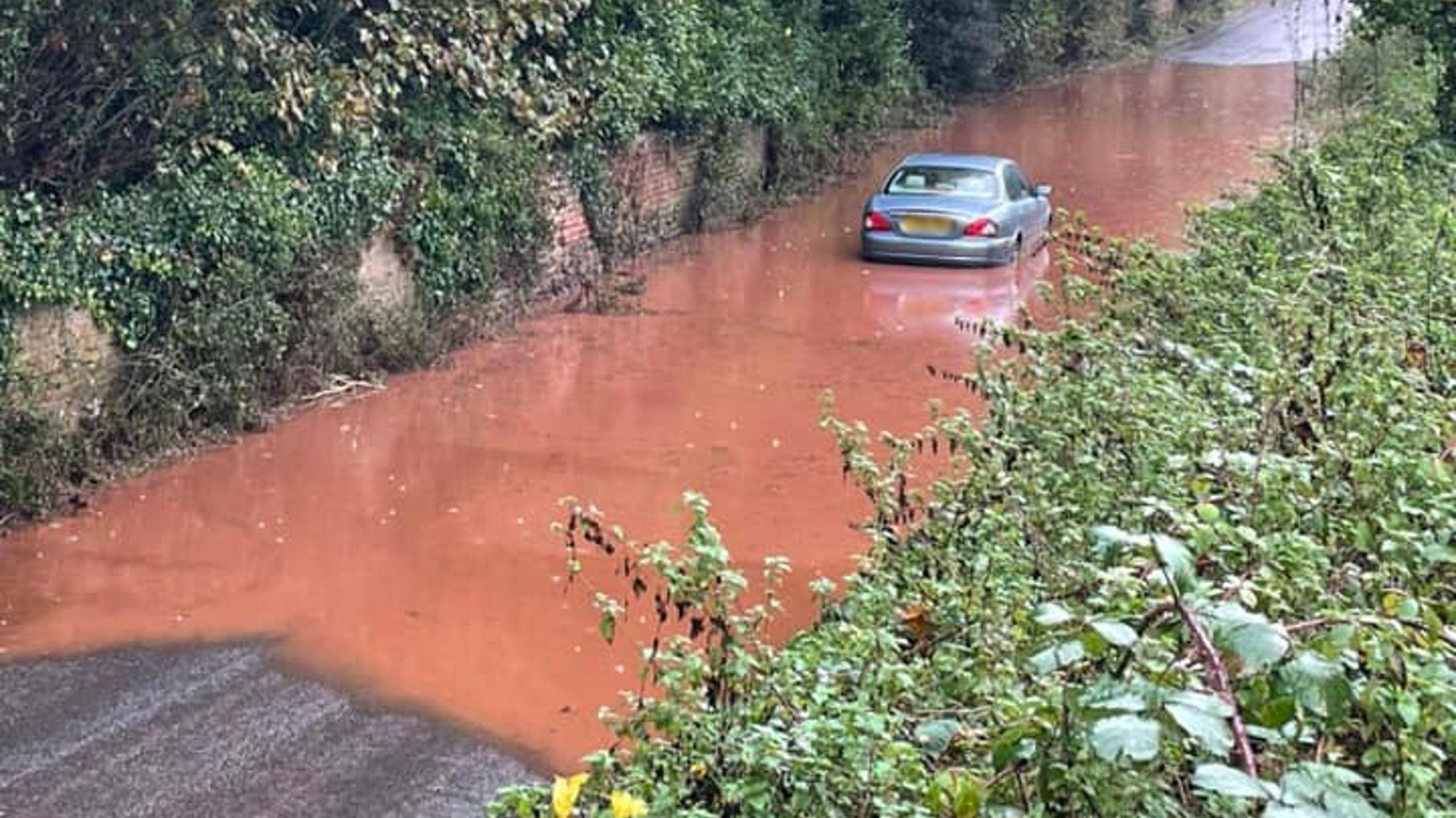 A silver vehicle stuck in flooded water on a road heading into Crediton from Sandford in Devon. The water is brown and there is a wall to the left of the road, with trees and brambles above and hanging down the road. There are also green brambles to right of the road.  