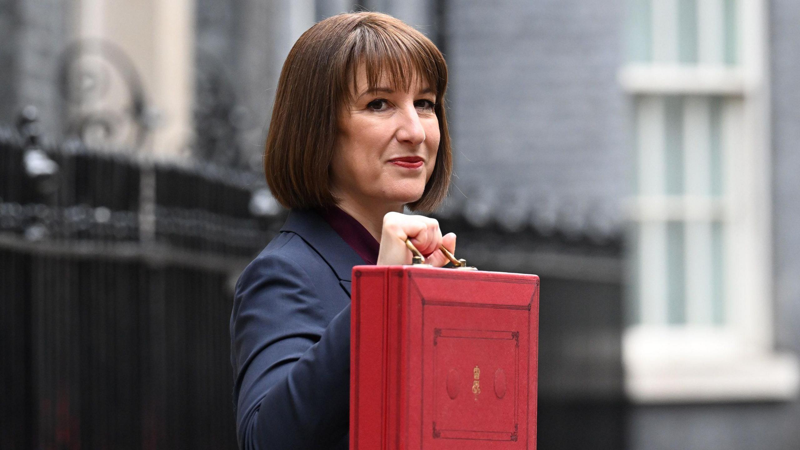 Chancellor of the Exchequer, Rachel Reeves, a woman with brown hair, is holding a red briefcase up as she poses for photographs in Downing Street.