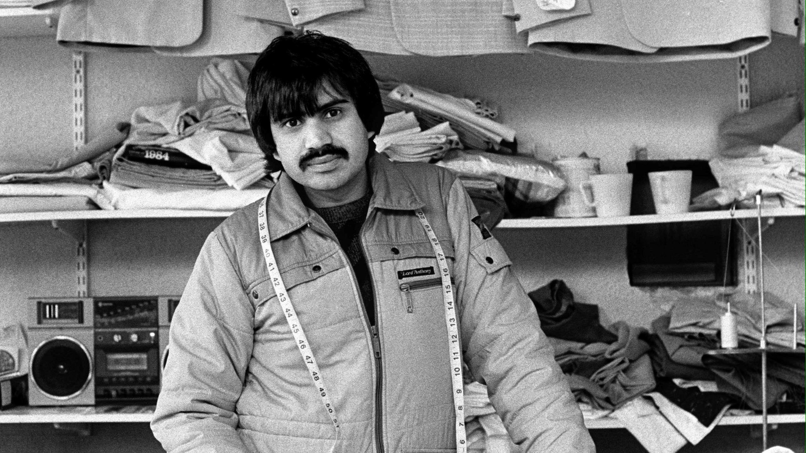 Black and white photograph of Barket Ali standing in his store with a tape measure around his neck, Nayyir Garments back in 1986