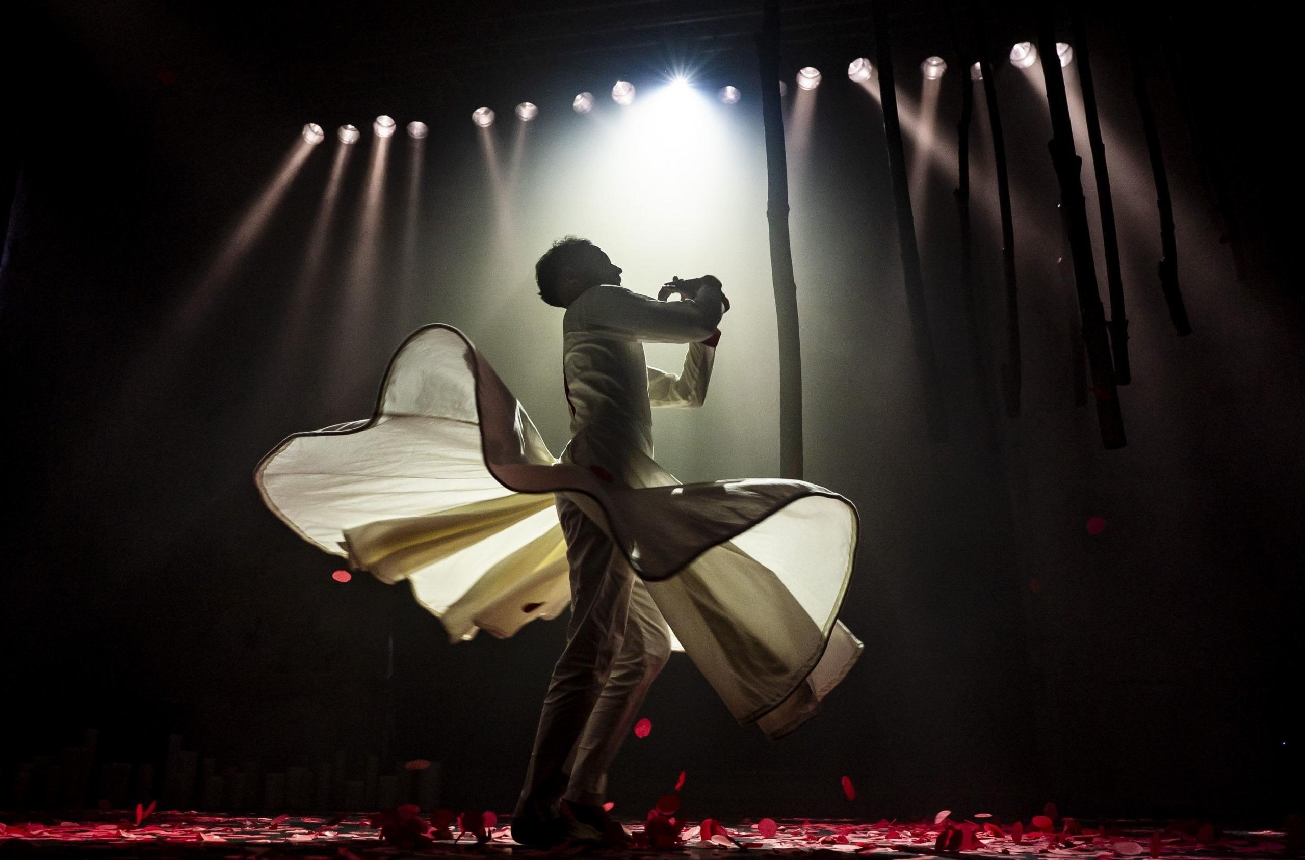 A man dances on stage at The Mount Without in Bristol. He is wearing a white outfit and is illuminated by multiple spotlights