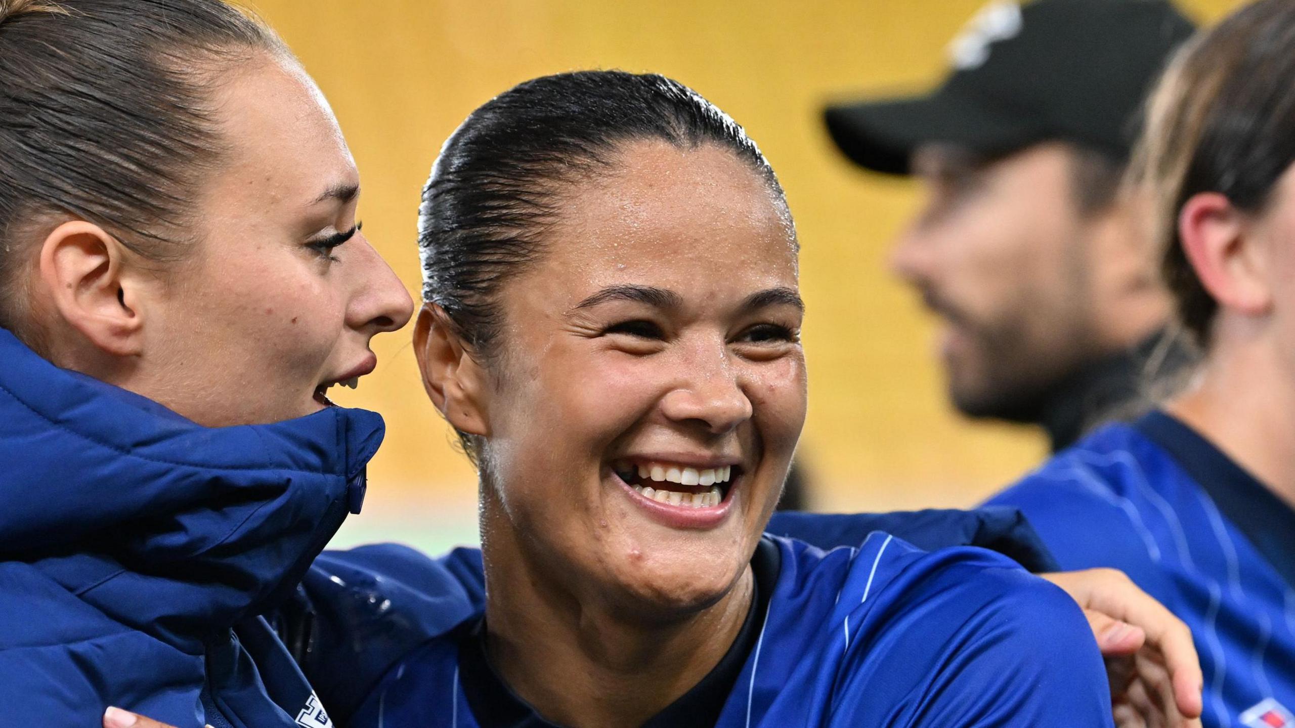 Natasha Thomas is pictured close up following a football game. She is smiling widely and facing away from the camera. Her dark hair is tied up behind her head. She is wearing a blue Ipswich Town shirt while a team mate hugs her from the side.