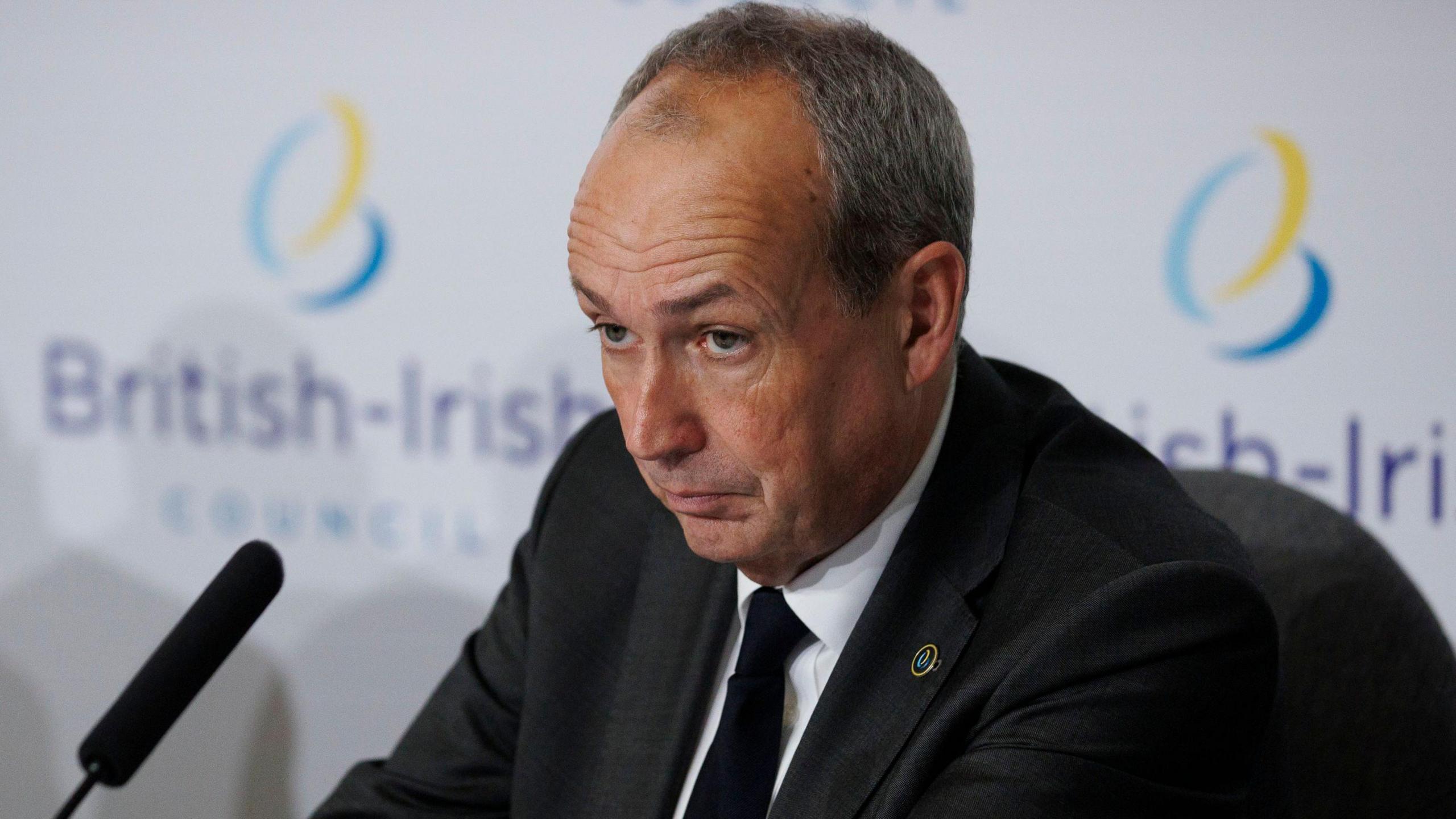 Deputy Lyndon Farnham, Chief Minister of Jersey, sits during a press conference at a British-Irish Council summit. He is wearing a suit with a black tie. A board behind him has two British-Irish Council logos on it. A microphone is in front of him.