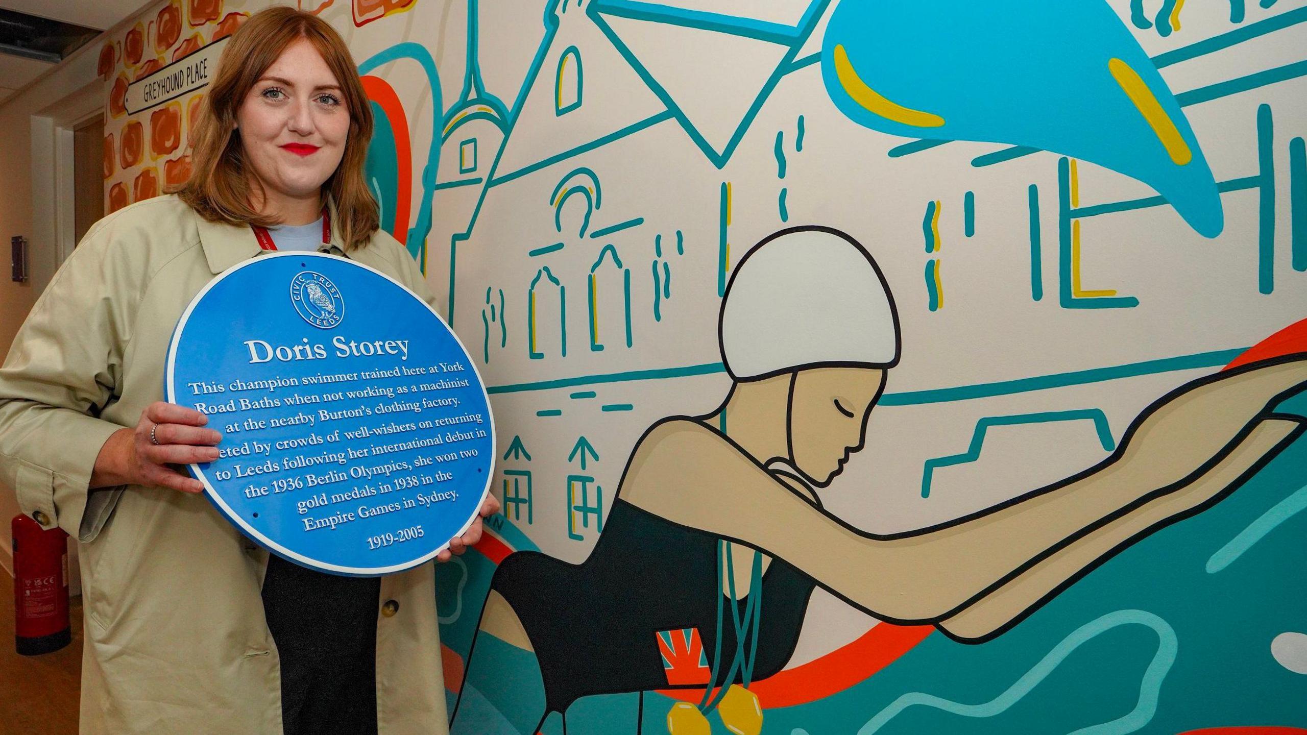 A woman with shoulder-length red hair holds a blue plaque while standing next to a mural showing a woman swimming with gold medals around her neck and a Great Britain flag on her swimsuit.