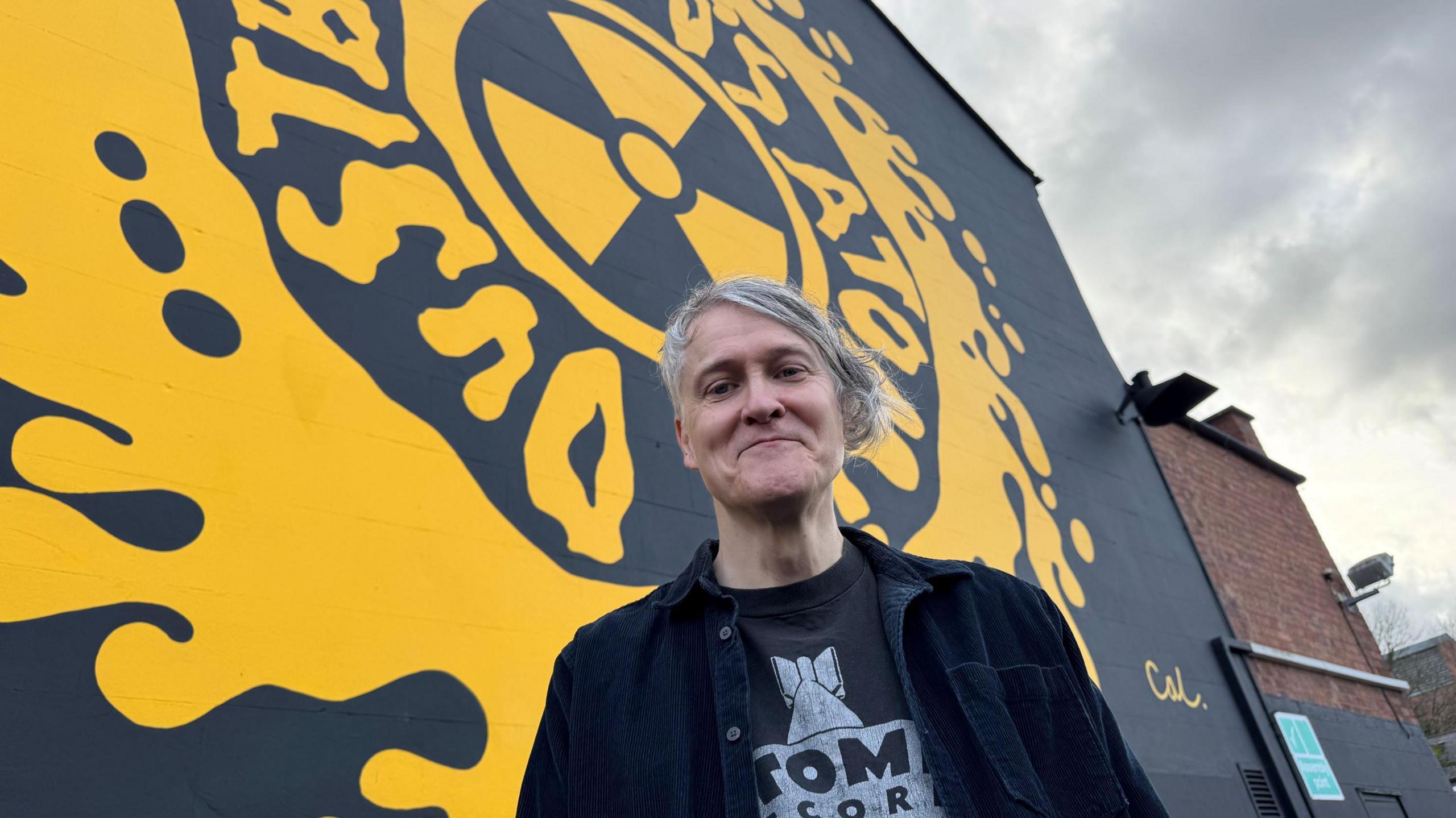 Jonn Penney  has grey hair and is smiling at the camera. He wears a black shirt and T-shirt with writing on. Behind him is a yellow and black mural. 