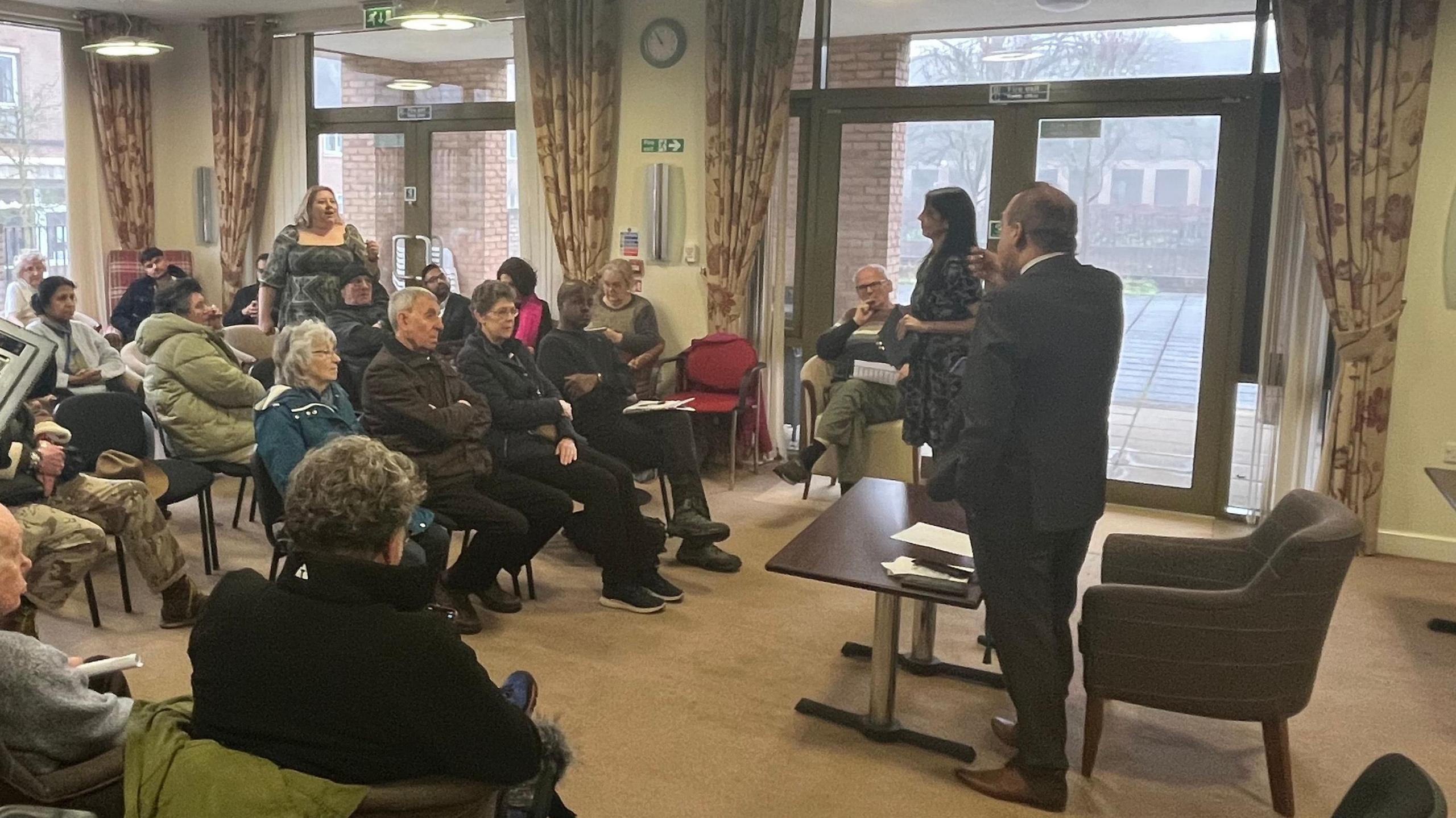 People sat in rows of chairs looking towards a table where a man is stood up.