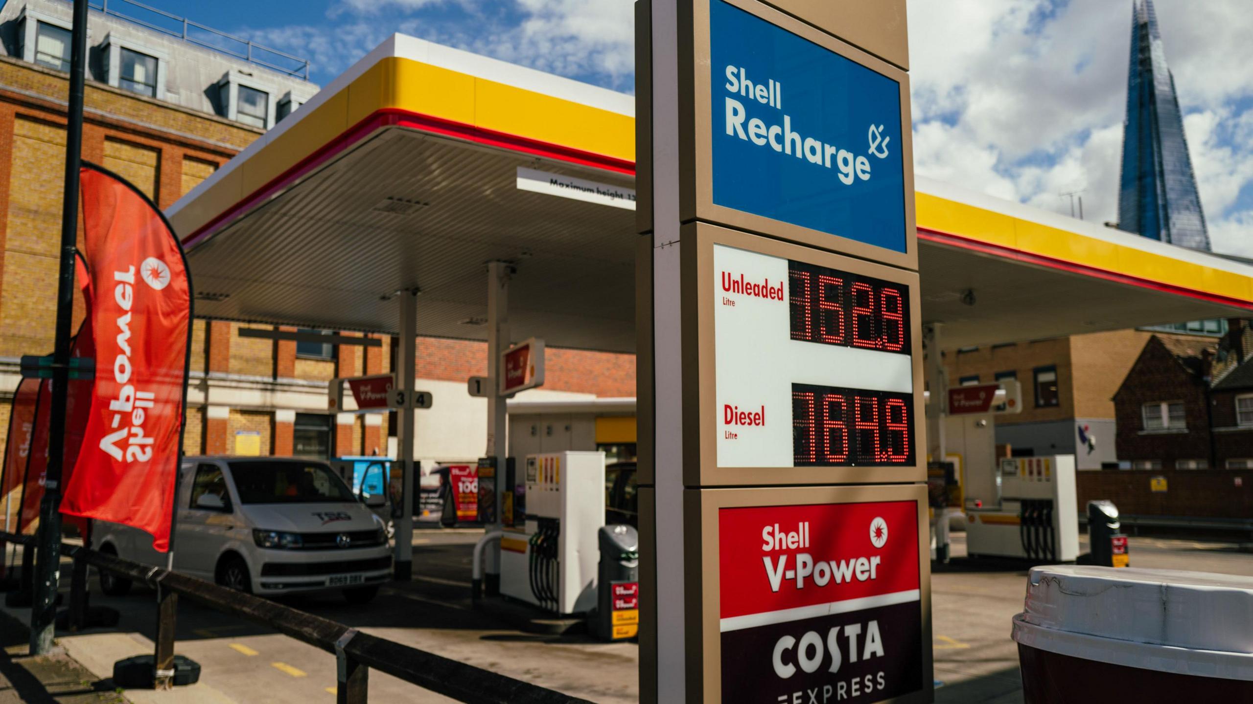 Shell petrol station court with a van beside a pump. A sign in the forefront shows the prices beside each fuel. 