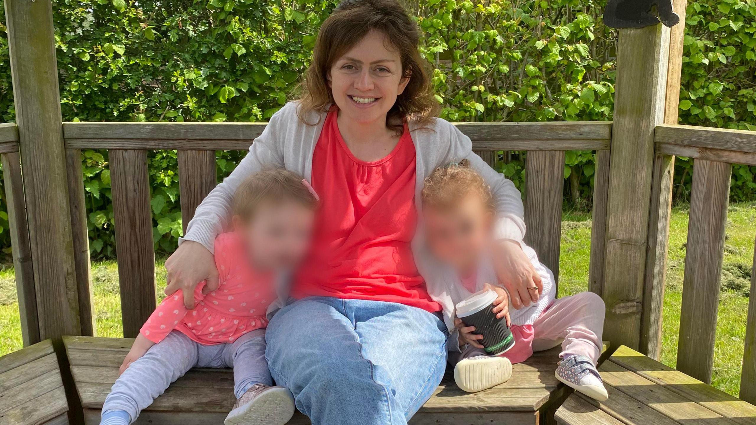 A woman sitting on a bench with her arms around two small children. She has brown hair and is wearing a pink t-shire, grey cardigan and blue jeans, and is smiling at the camera. The children both have blonde hair and their faces have been blurred out. One is holding a takeaway coffee cup.