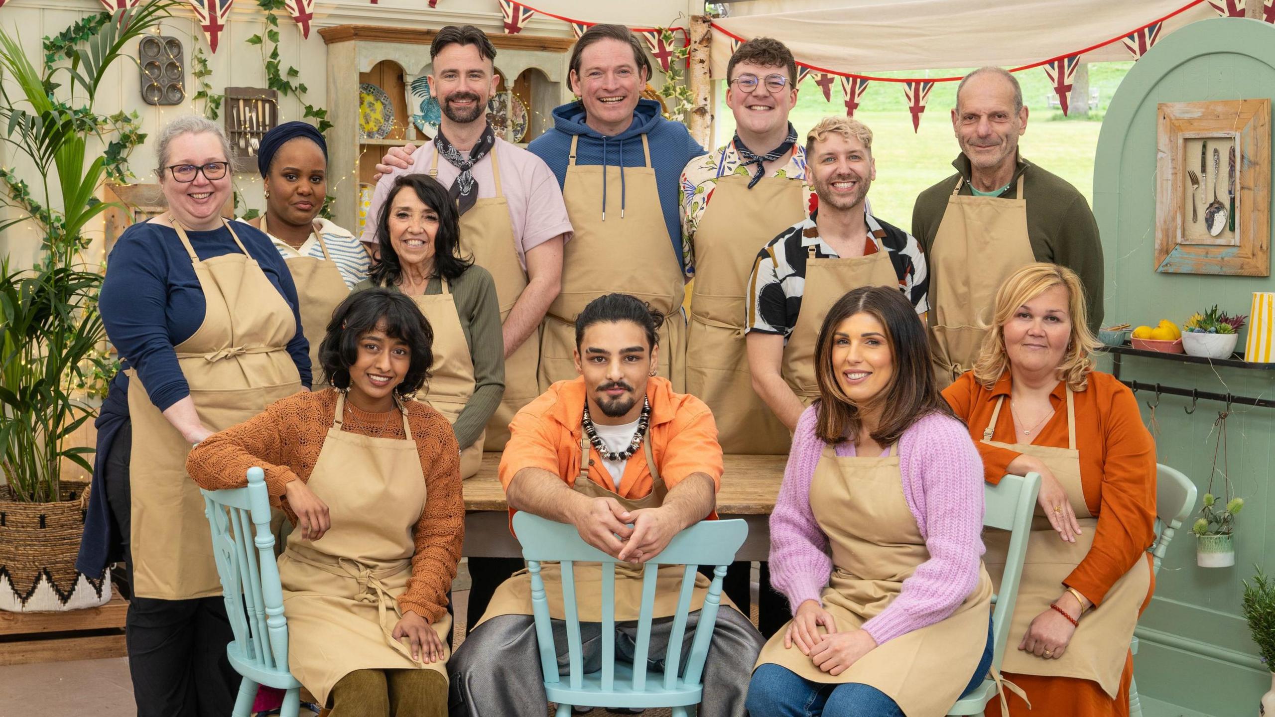 Twelve people in a group wearing brown aprons in the Great British Bake Off tent. 