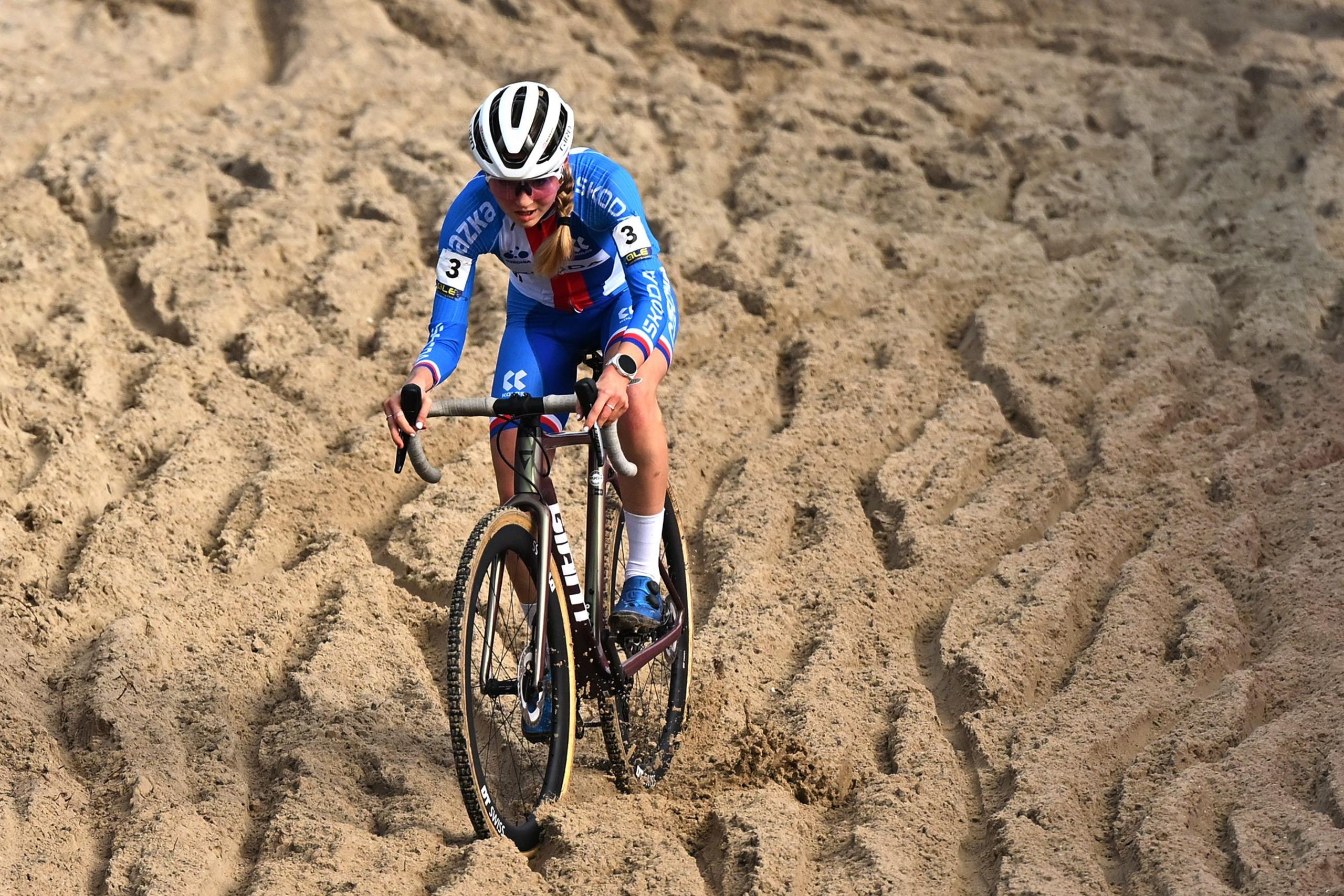 A cyclist wearing a blue racing kit and a white helmet rides a cyclocross bike through deep sand, with visible tire tracks across the sandy surface.