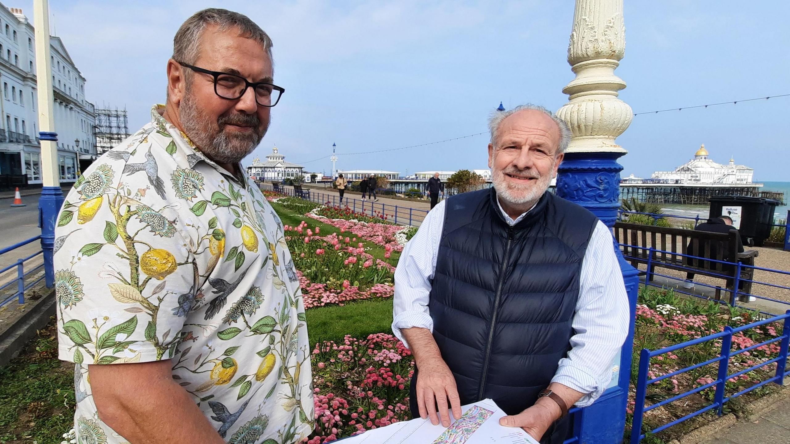 Councillor Jim Murray poses for a photograph with landscape and garden designer Peter Thurman. Councillor Murray is wear a white, short-sleeved shirt featuring a design of yellow lemons on branches. Mr Thurman is wearing a navy gilet over a light blue shirt with rolled-up sleeves. They are standing in from of Eastbourne Carpet Gardens, blooming with pink plants. The sky is clear blue and Eastbourne Pier is visible in the background.