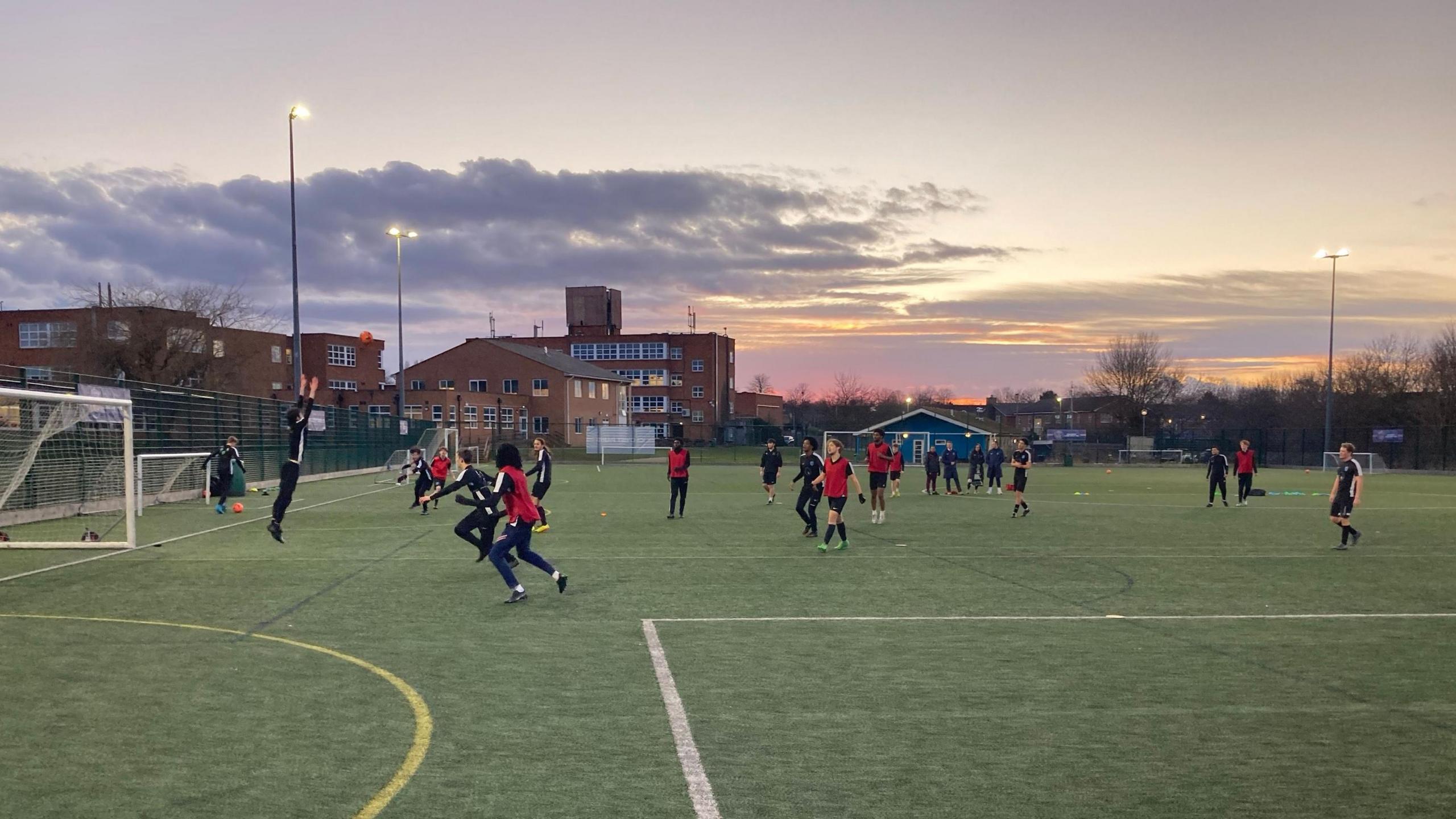 Youngsters on a school astro pitch playing football. Buidlings visible in the background and floodlights on.