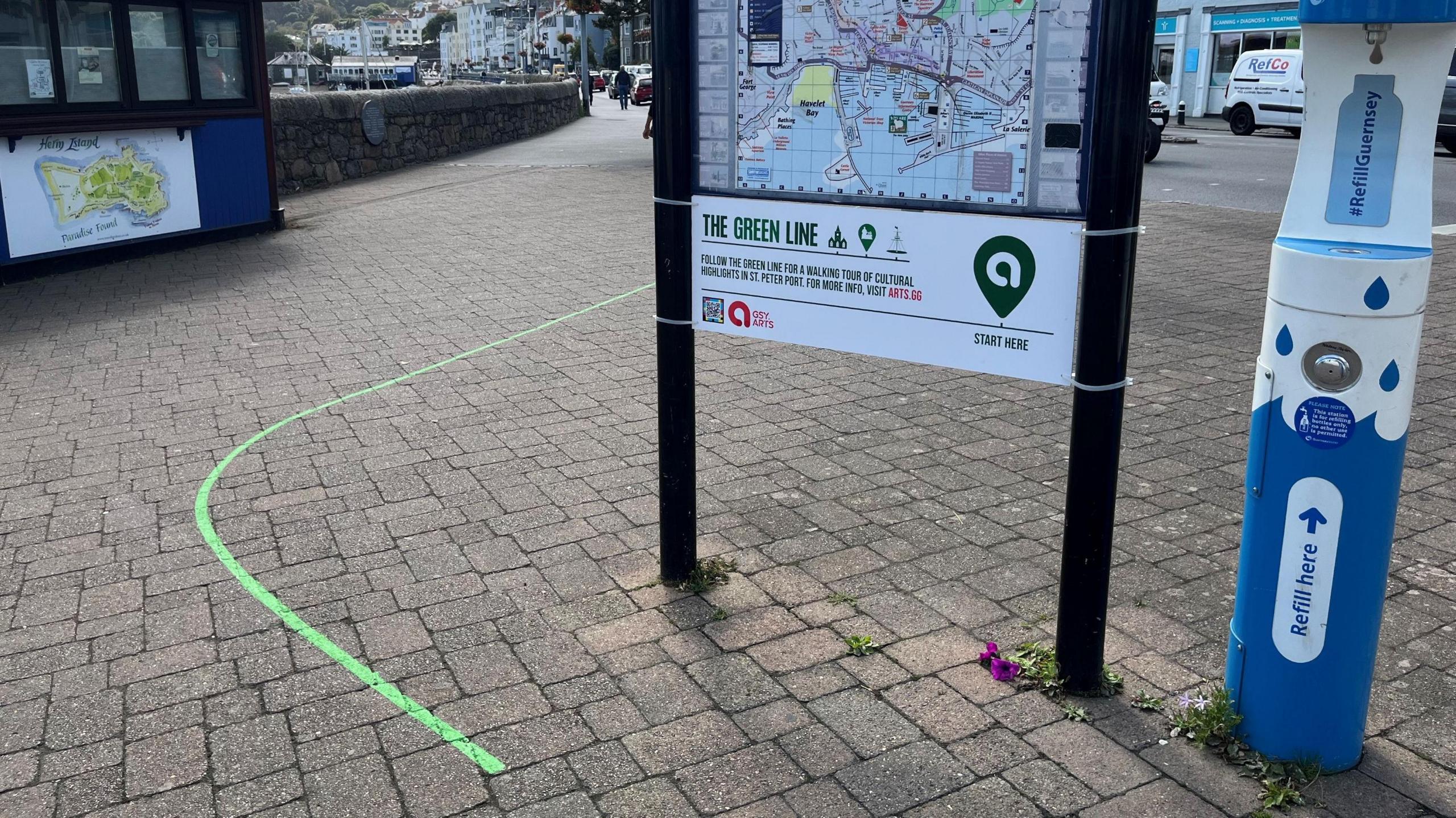 A green line painted on the ground starts by a sign showing St Peter Port and information about the green line. It stands next to a water fountain. In the background the Travel Trident Kiosk, a pavement, traffic and St Peter Port seafront can be seen.