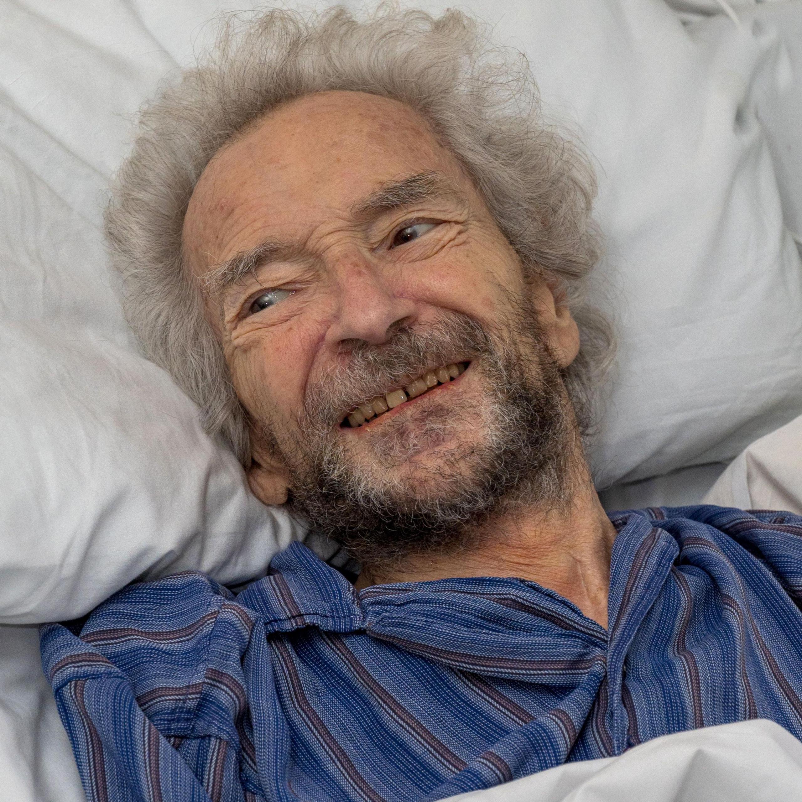 Alan Cohen, 81, lying in bed, smiling, while looking off camera to the left