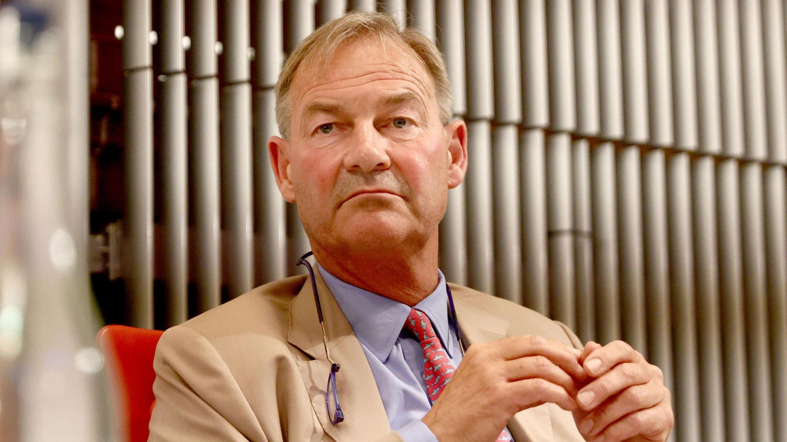 Rupert Lowe with short blond hair wearing a cream jacket, light purple shirt and red patterned tie.