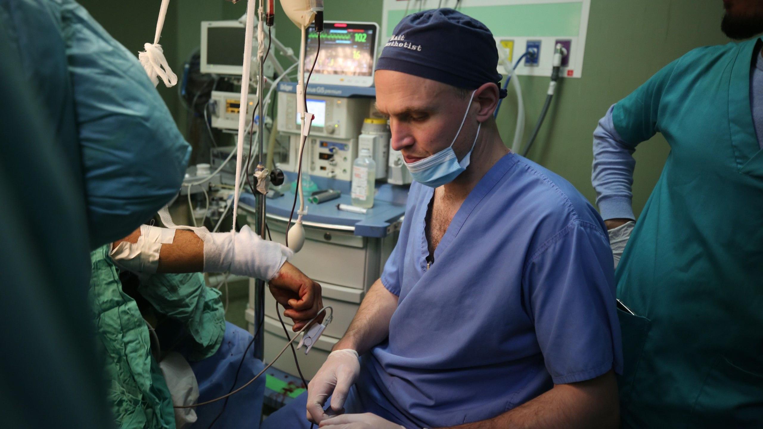 Dr Matt Newport sits next to a patient during surgery in a field hospital in Gaza, wearing his blue surgical scrubs and surgical cap. The bandaged arm of a patient is just visible next to him.