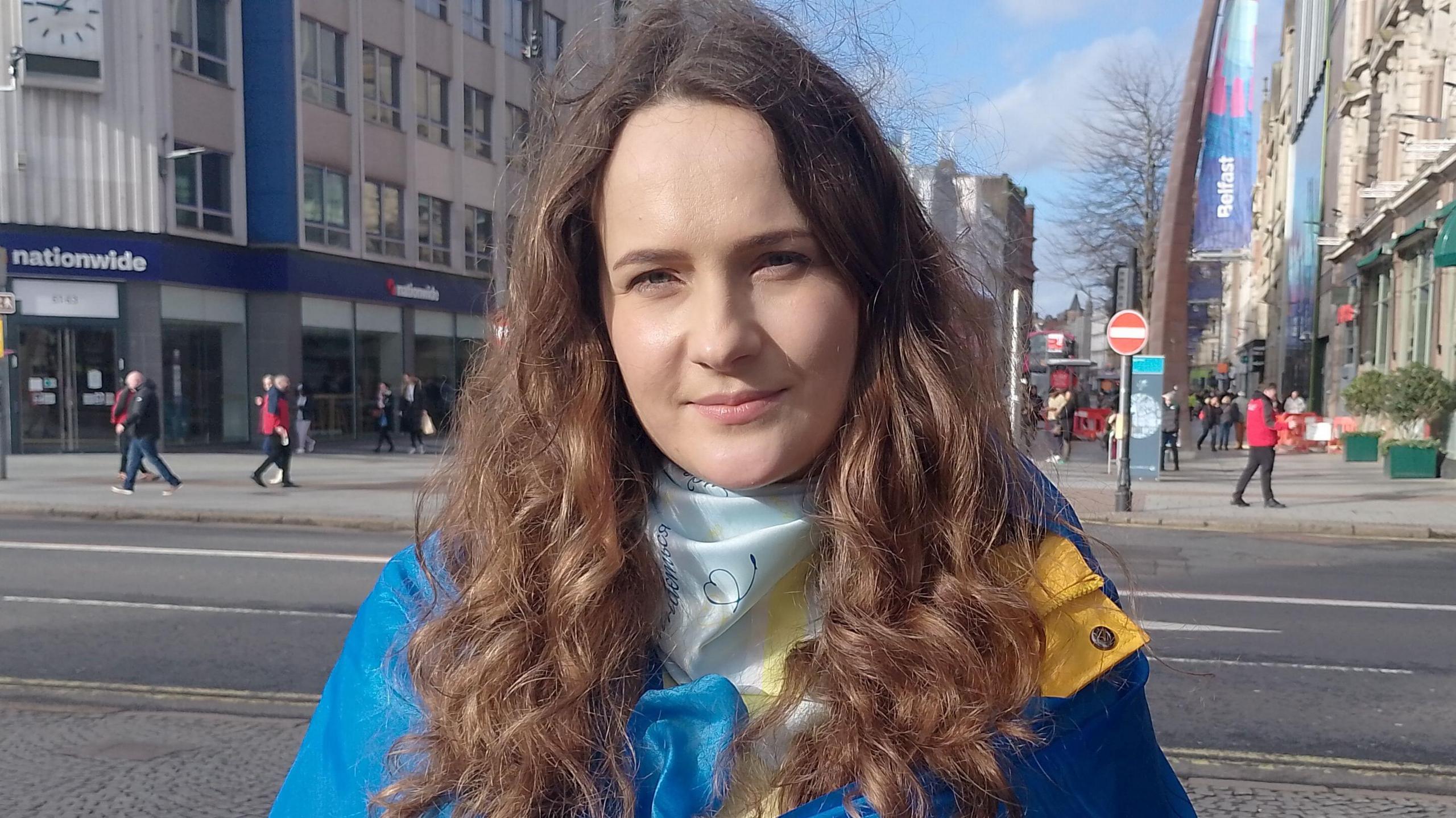 A woman looks into the camera. Her long curly hair is brown. She's wearing the Ukranian flag draped over her shoulders. A city centre is behind her.