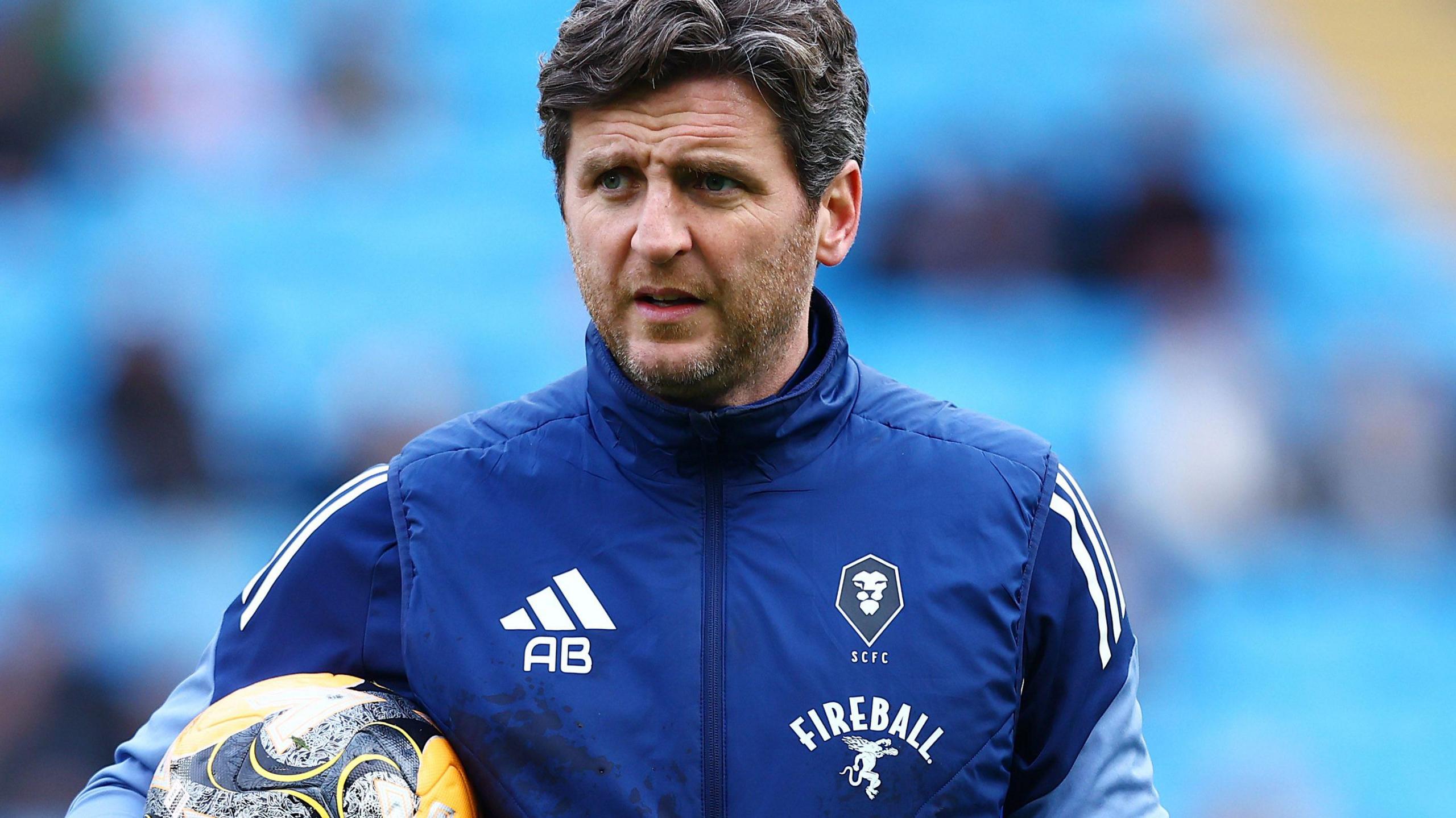 Alex Bruce during his time as Salford City assistant manager wearing a blue club jacket and holding a football under his right arm.