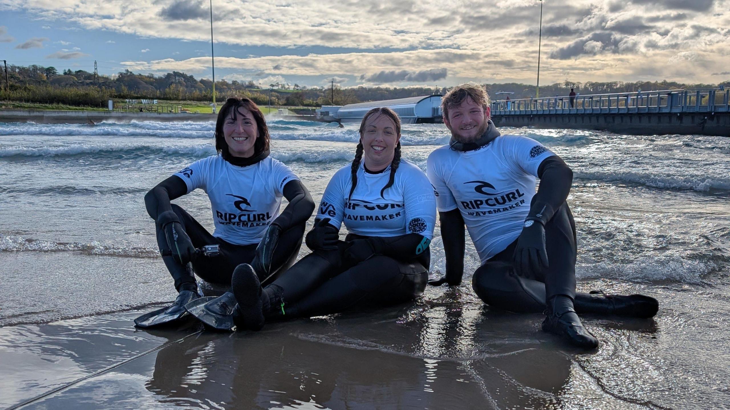 Three people sit on the shore of an artificial beach in Bristol. In the middle is Kay Millar, who is sitting with her legs outstretched in front of her. Her hair is tied in tow ponytails. On her right is her coach Hannah Mattison, who has shoulder-length brown hair. On Kay's left is her coach Ryan Trott who has short brown hair. They are all wearing black wetsuits with white Rip-Curl rash vests on top.