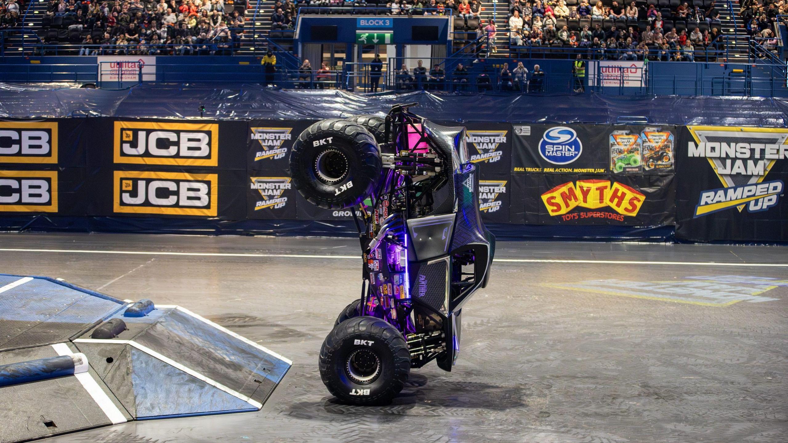A monster truck driving upright on its two front wheels along the floor of an arena next to a large ramp. There are fans in the stand behind it. The truck is black and purple with pink lights on it