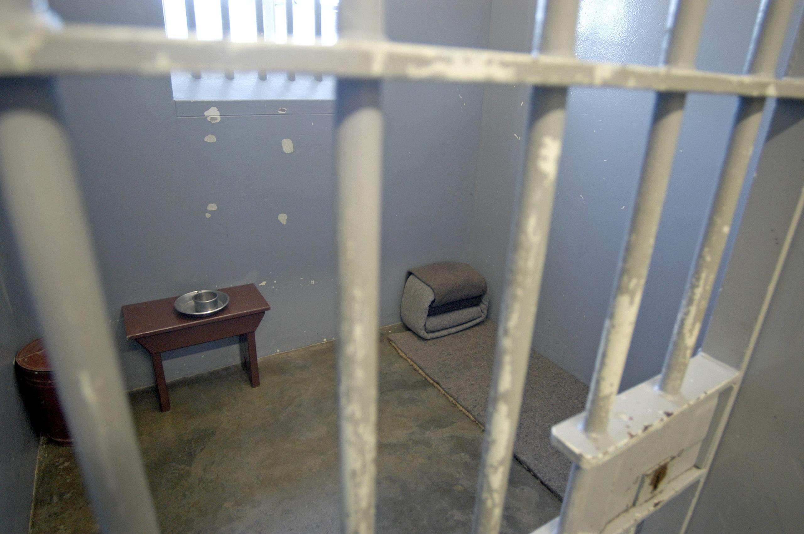 a picture of an empty cell with a blanket bed on the ground and a small table with a metal plate and bowl. There are bars across the window.