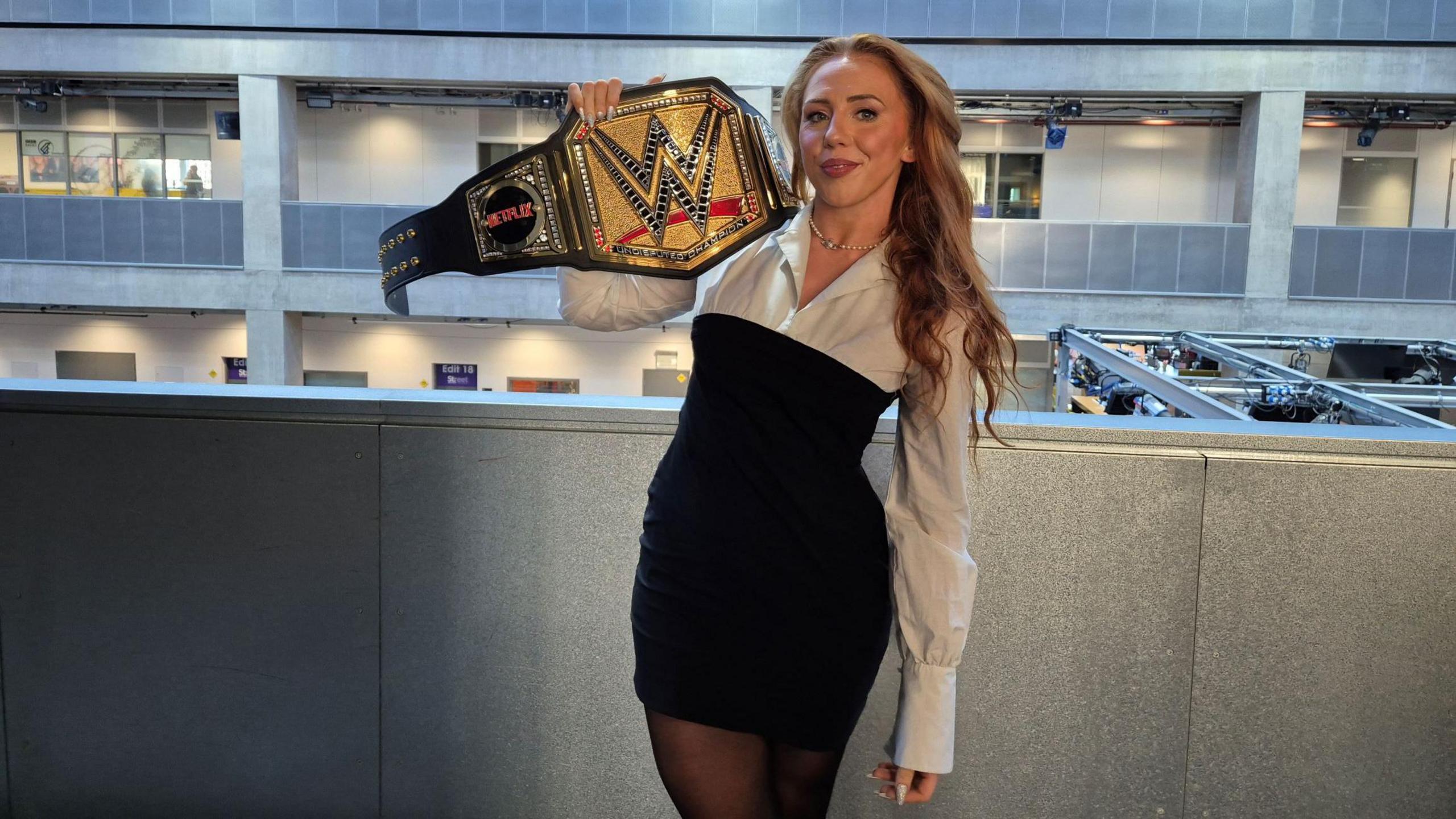 Isla Dawn, wearing a black and white dress, holds a WWE title up while standing in an office building. She has long red hair and is smiling