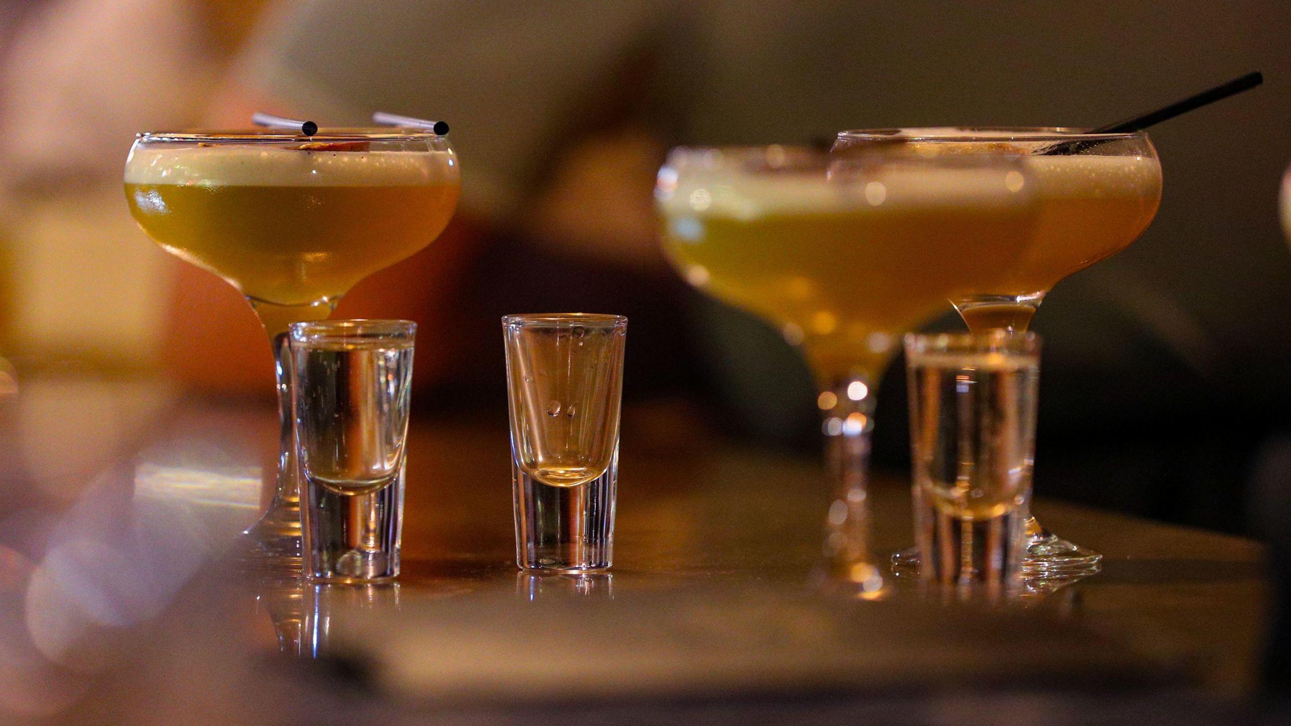 Three full cocktail glasses and three full shot glasses on a bar. The cocktail is orange in colour, the shot is a clear liquid. The background is blurred out of focus.