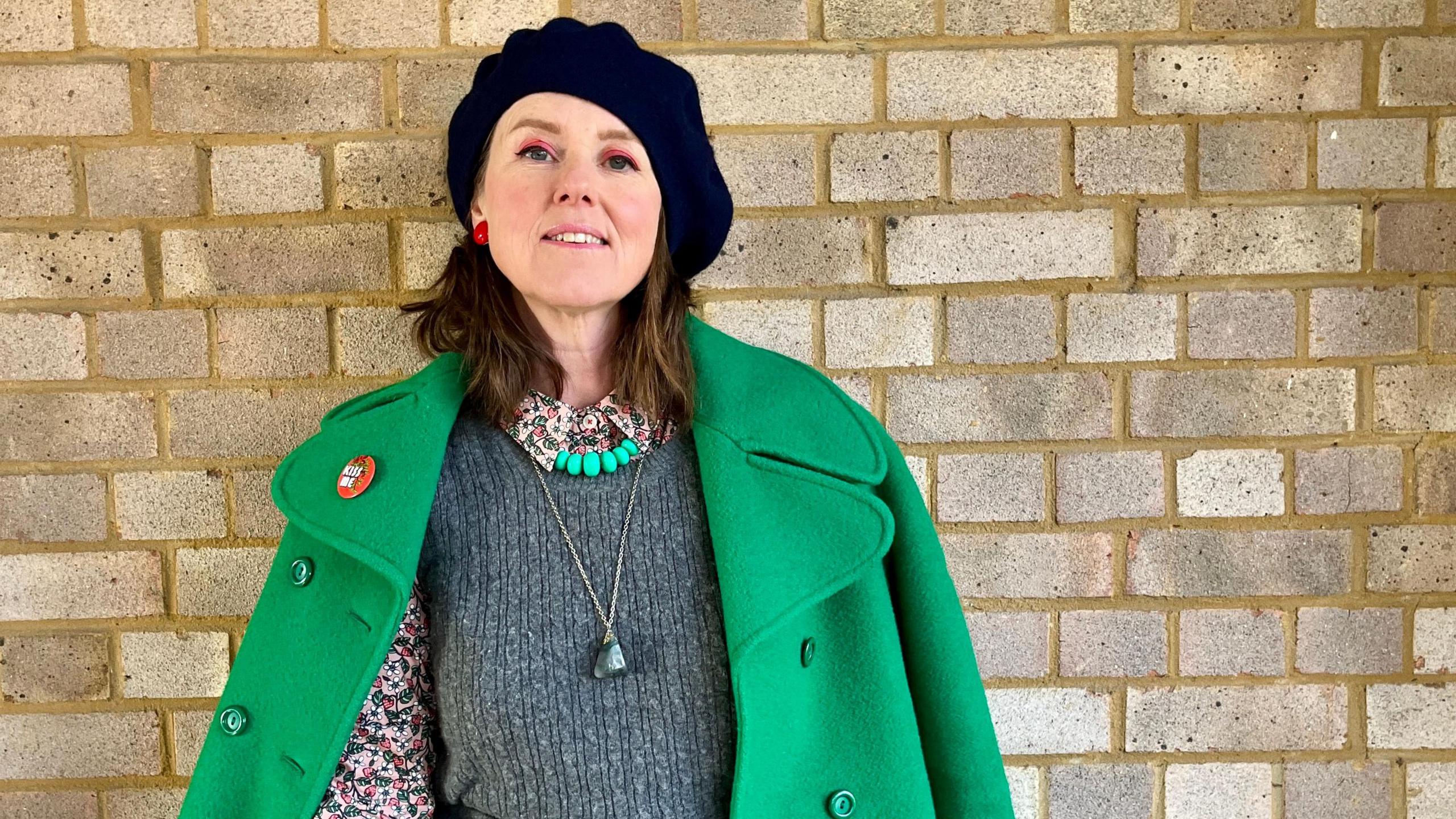 Caroline Jones in 2025 has brown shoulder-length hair and is wearing a navy blue beret. She also wears a floral blouse under a grey tank top and a bright green coat. She wears a necklace with a green stone.