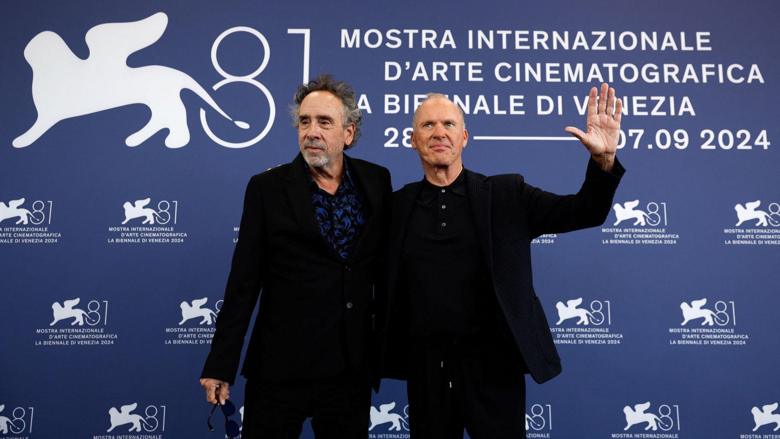 Tim Burton and Michael Keaton on the red carpet at the 81st Venice Film Festival both men are wearing black and Keaton has one arm around Burton and the other raised in the air waving