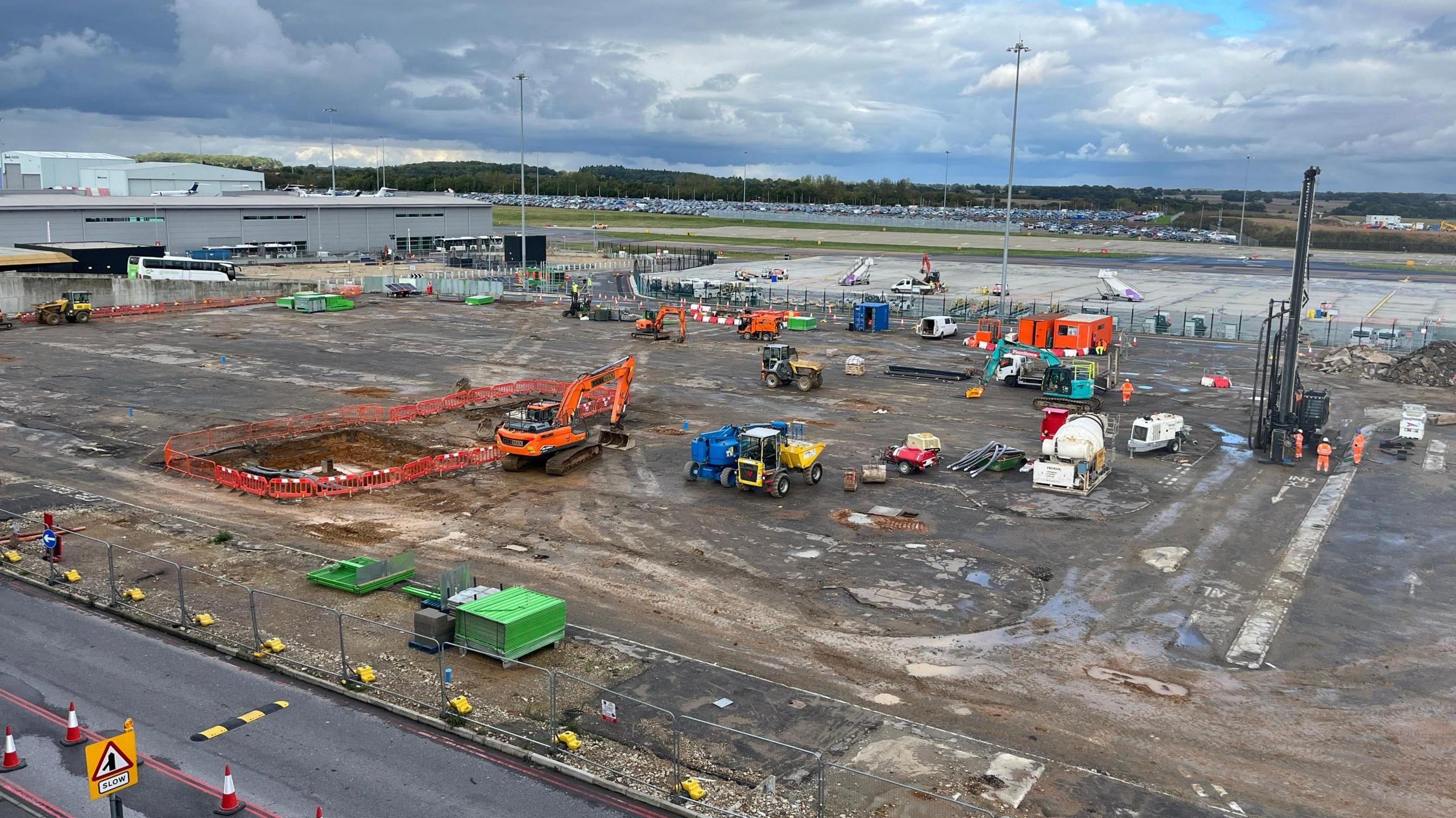 The site where the car park once was. It has many construction vehicles and workers on it. The site lies next to a small runway and it is mostly covered in mud.