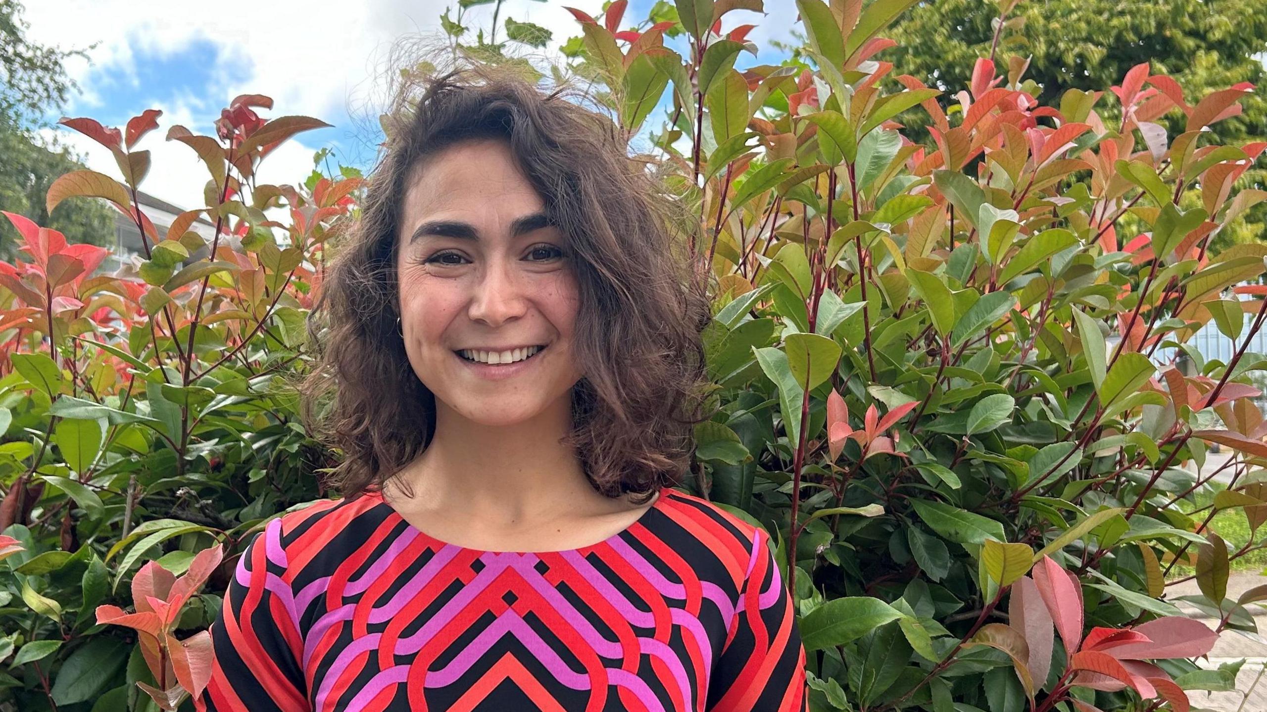 A smiling Parwana Fayyaz looks directly at the camera wearing a black top with a pink and red pattern. She is standing in front of a green bush. 