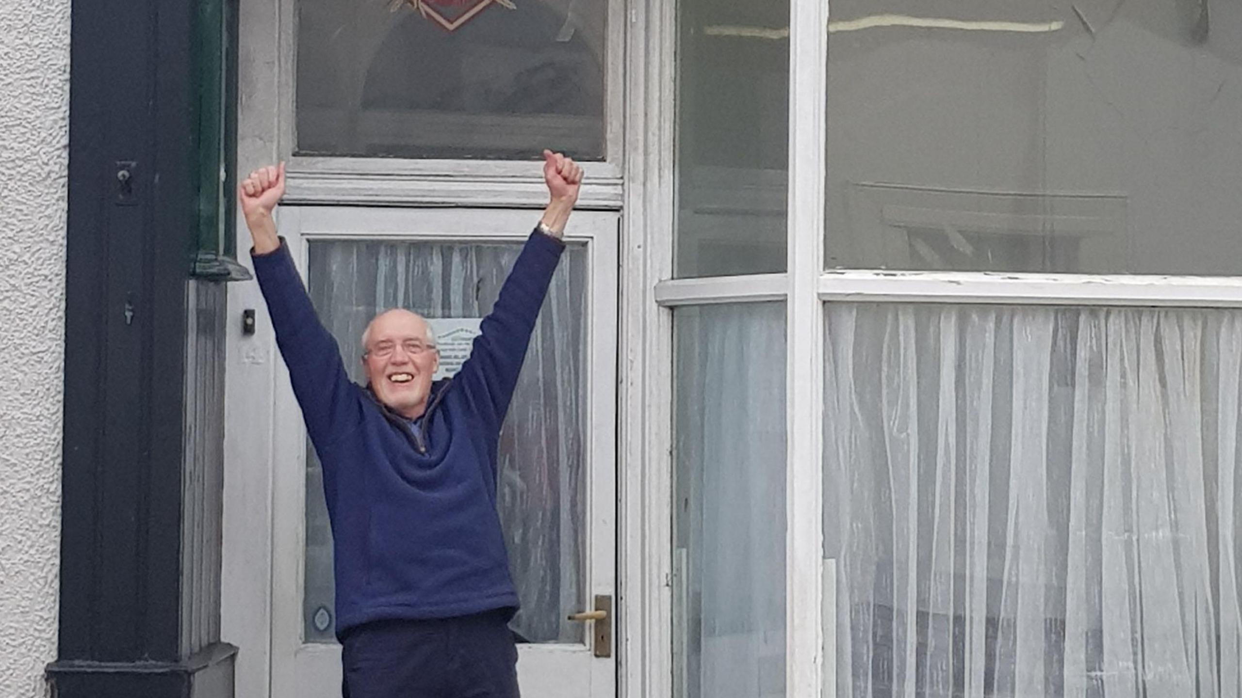 Charlie Lambert celebrating outside Norman Nicholson's house after the property was bought in February. Mr Lambert is smiling and his hands are up in the air. He is wearing a navy blue sweatshirt and has glasses. The house has large front windows with white panes.
