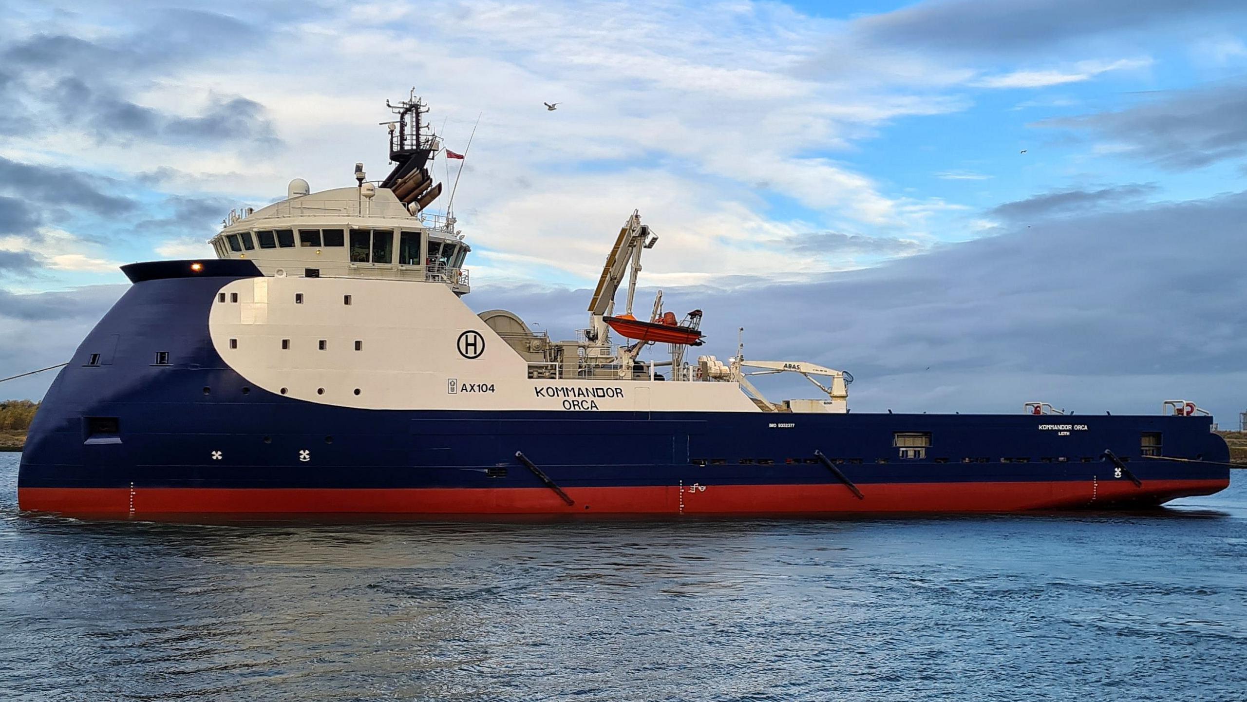 The blue, white and red-painted Kommandor Orca at sea. A deck crane is visible halfway along the ship in front of the cargo deck.