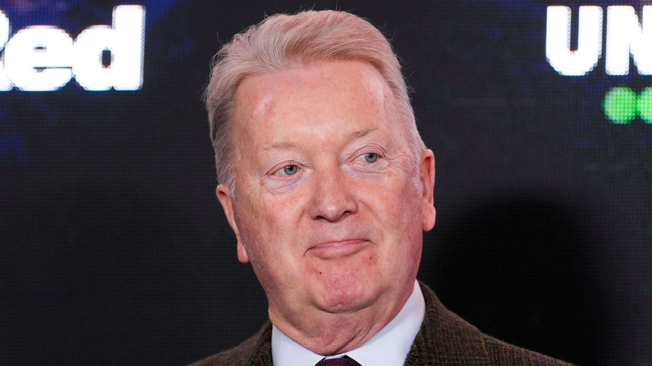 Promoter Frank Warren looks away at the camera as he slightly smiles. He has short grey hair. He wears a brown suit jacket, white shirt and tie. He is standing in front of a large screen with some brand names on it that cannot be read. 